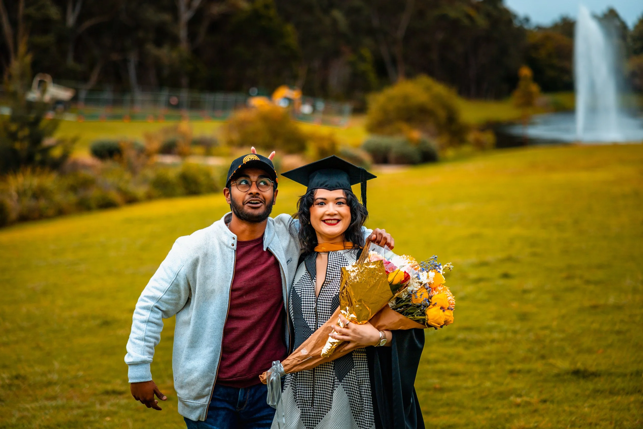 A woman in a graduation cap and gown holding a bouquet of flowers, standing with a man in casual clothes, both smiling and celebrating outdoors near a waterfall.