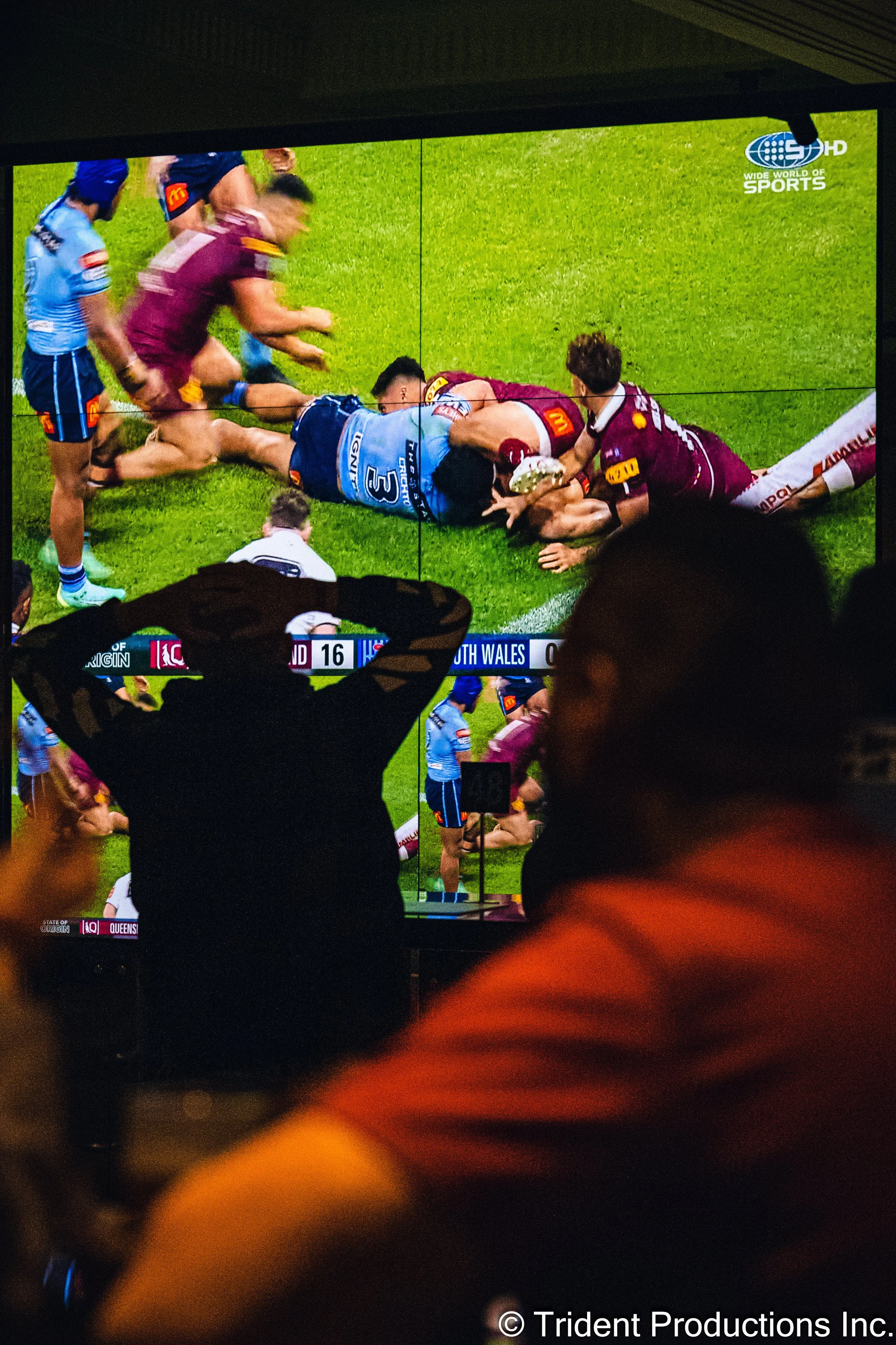 People watching a rugby game on a large screen, with one person in the foreground raising their arms, indoors.