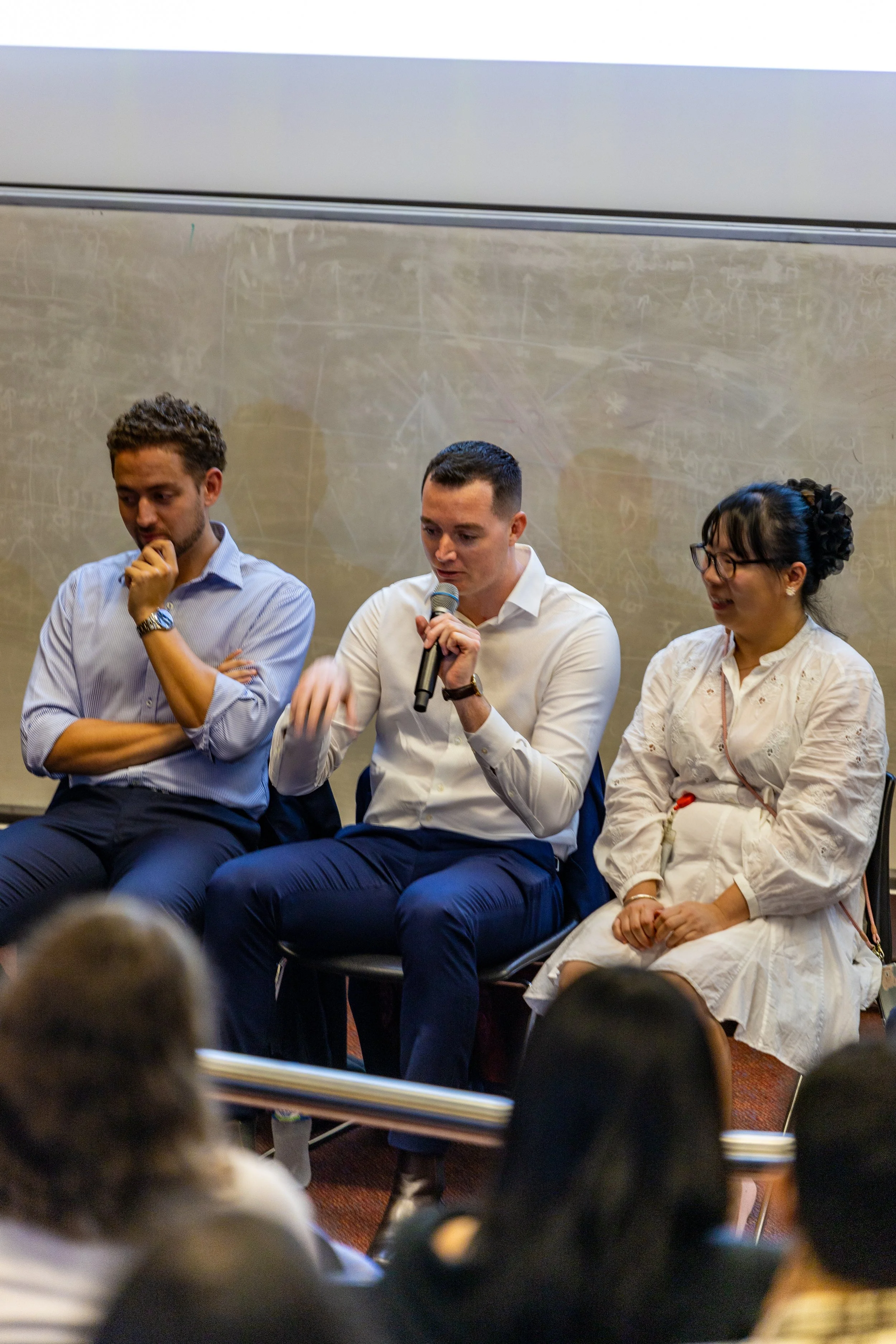 Three individuals sitting on chairs in a panel discussion or presentation setting. The middle person is speaking into a microphone. Behind them is a chalkboard with writing.