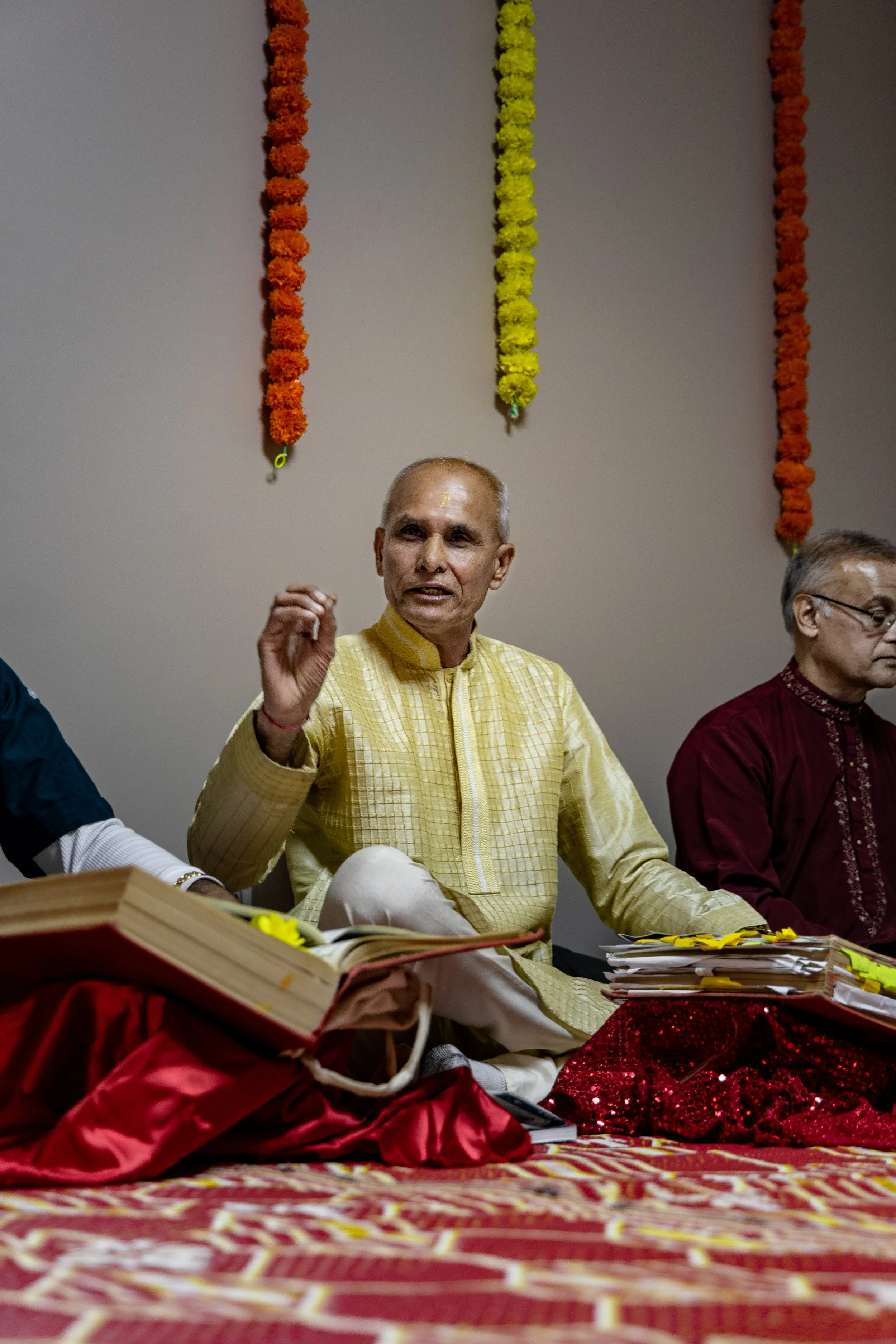Man in yellow traditional Indian attire speaking during a cultural or religious event, seated on a decorated platform with books and papers in front of him, floral garlands hanging behind.