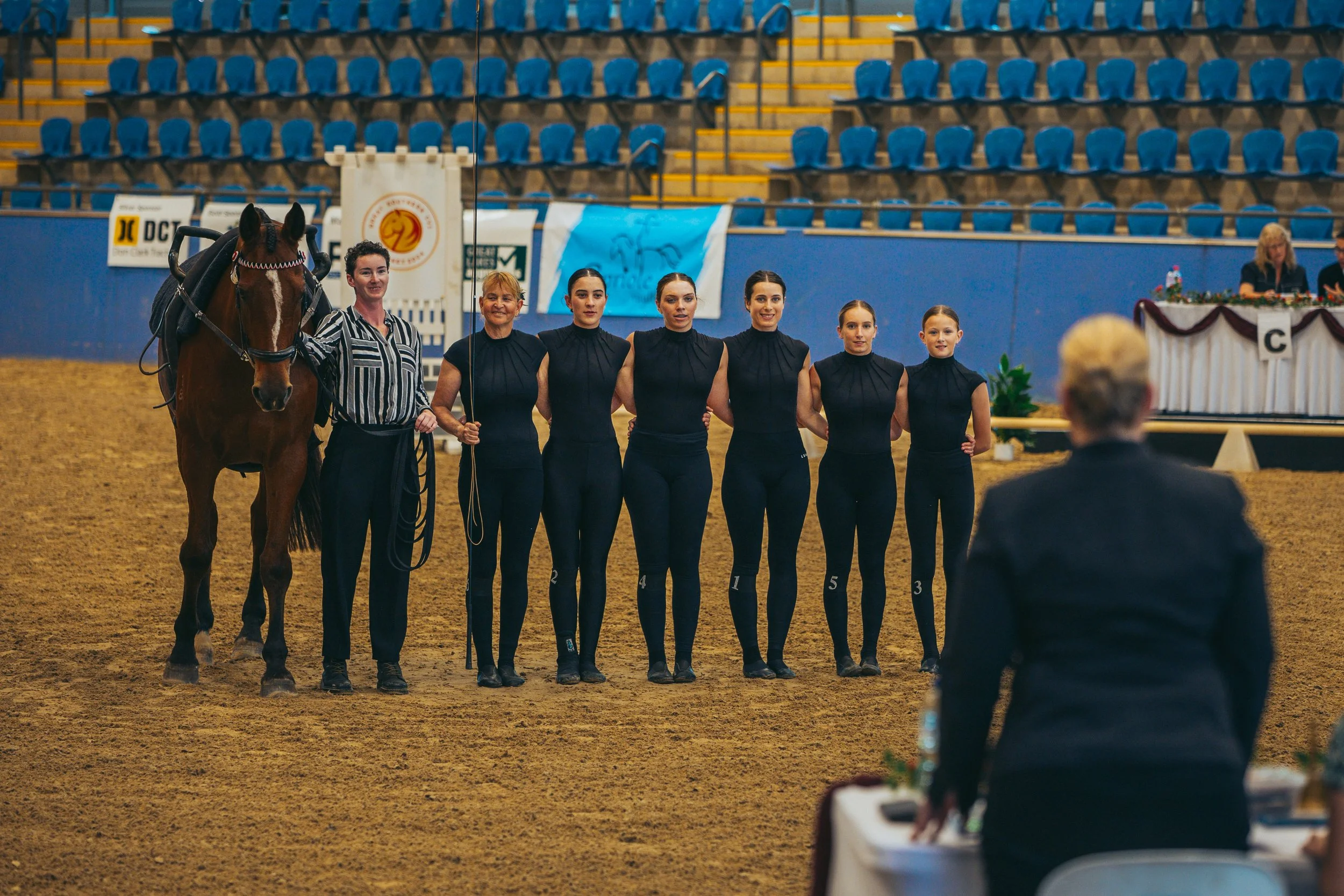 A group of six women in black riding outfits standing with a woman holding a horse and a person sitting at a table in an indoor arena with empty seats in the background.