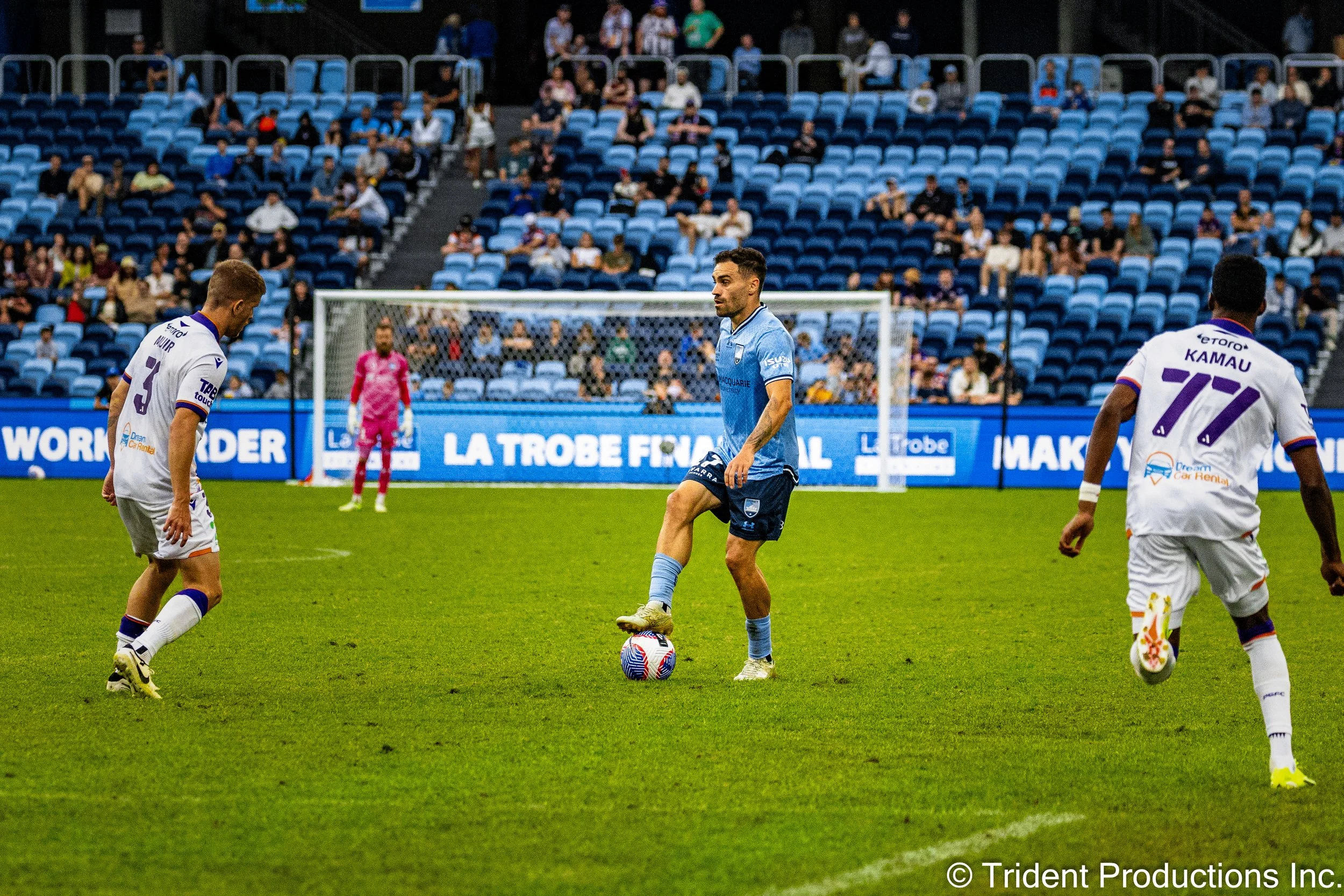Soccer players on the field, one in a blue uniform controlling the ball, two in white uniforms approaching, goalkeeper in pink watching, stadium with blue seats and spectators.