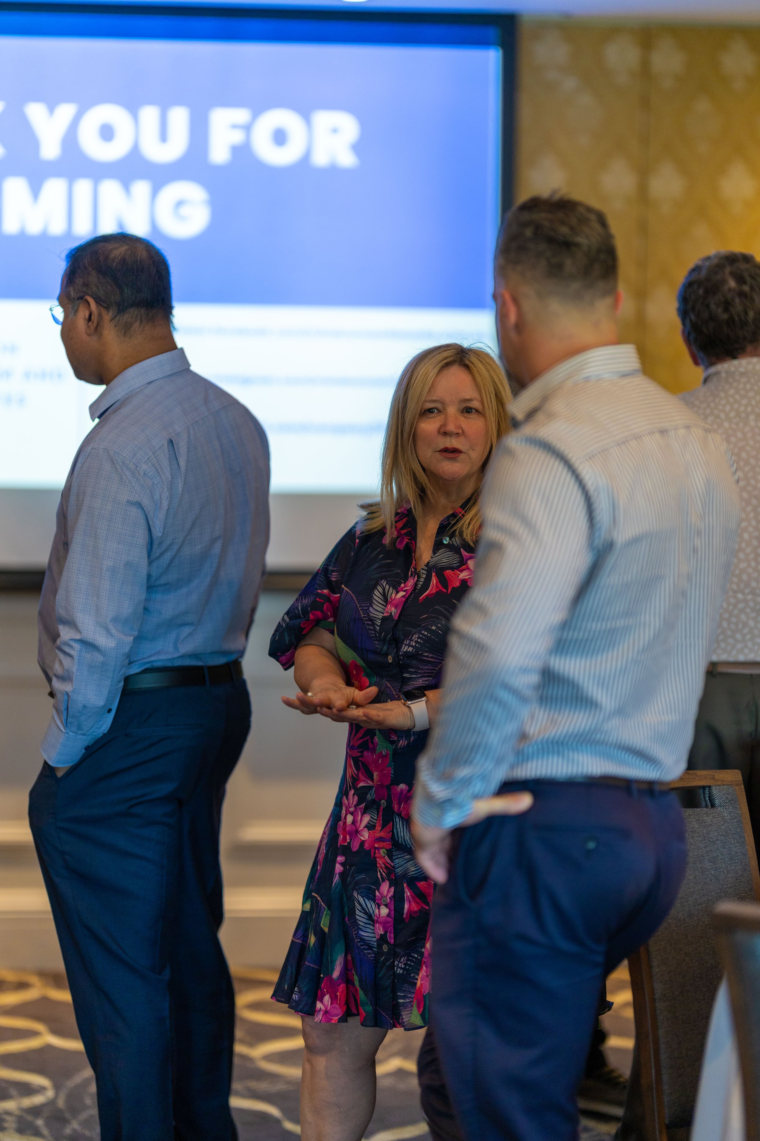 Group of professionals conversing at a conference with a large screen in the background that displays the words "Thank you for coming.".