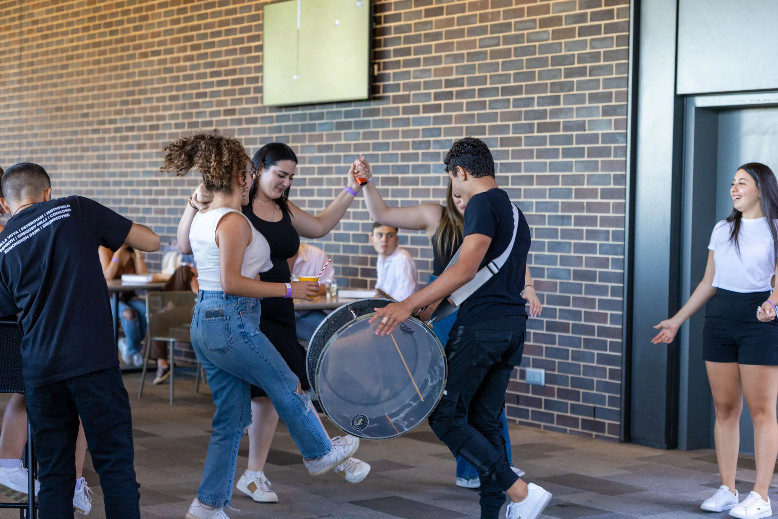 Group of young people dancing and having fun indoors with a brick wall background, some holding drinks, others enjoying dancing.