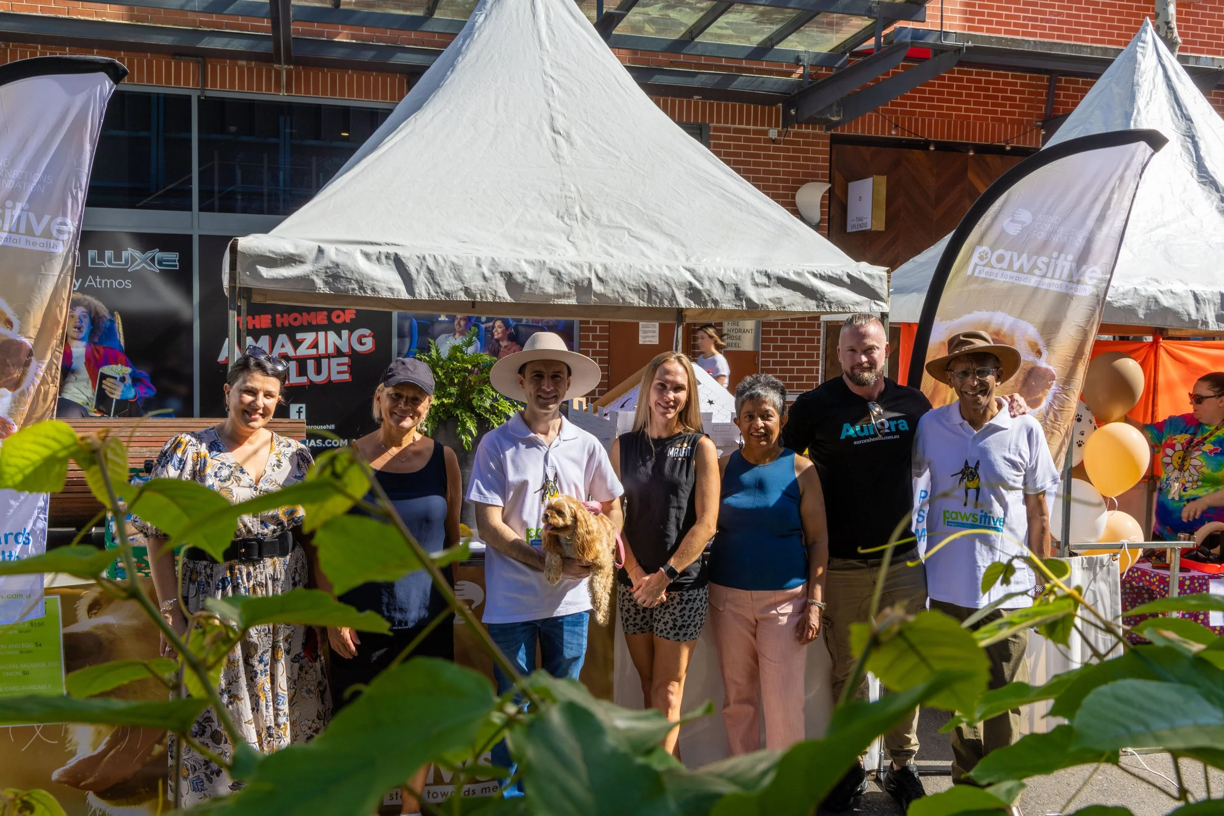Group of seven people posing in front of a booth at an outdoor event, with tents and banners, and some greenery in the foreground.