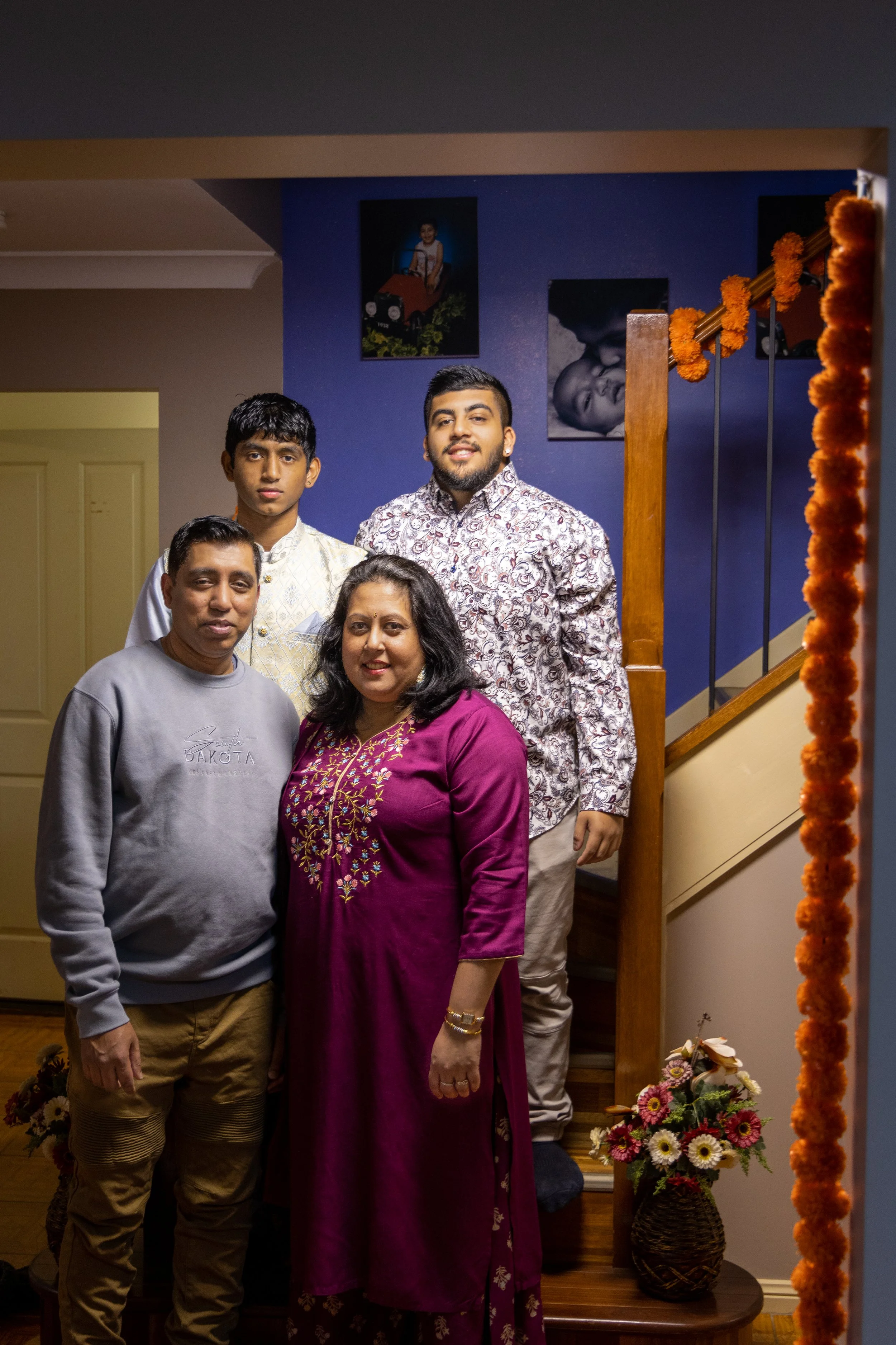 Family portrait of five people standing on a staircase inside a home, with framed photos on the wall behind them and a flower arrangement at the bottom of the stairs.