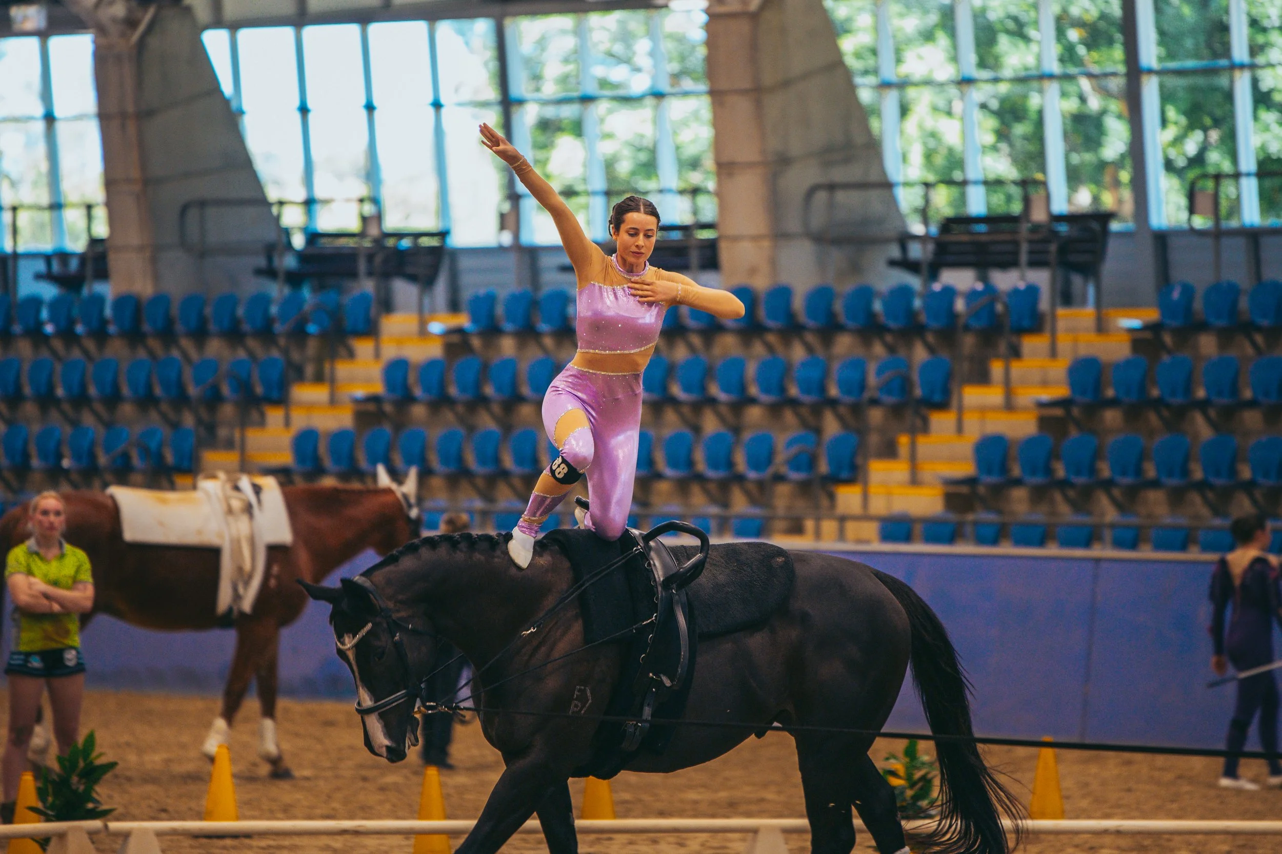 A female gymnast in a pink outfit performing a balancing act on a black horse in an indoor arena. She is standing on the horse's back with one leg bent and the other extended forward, arms raised and one hand placed on her chest.