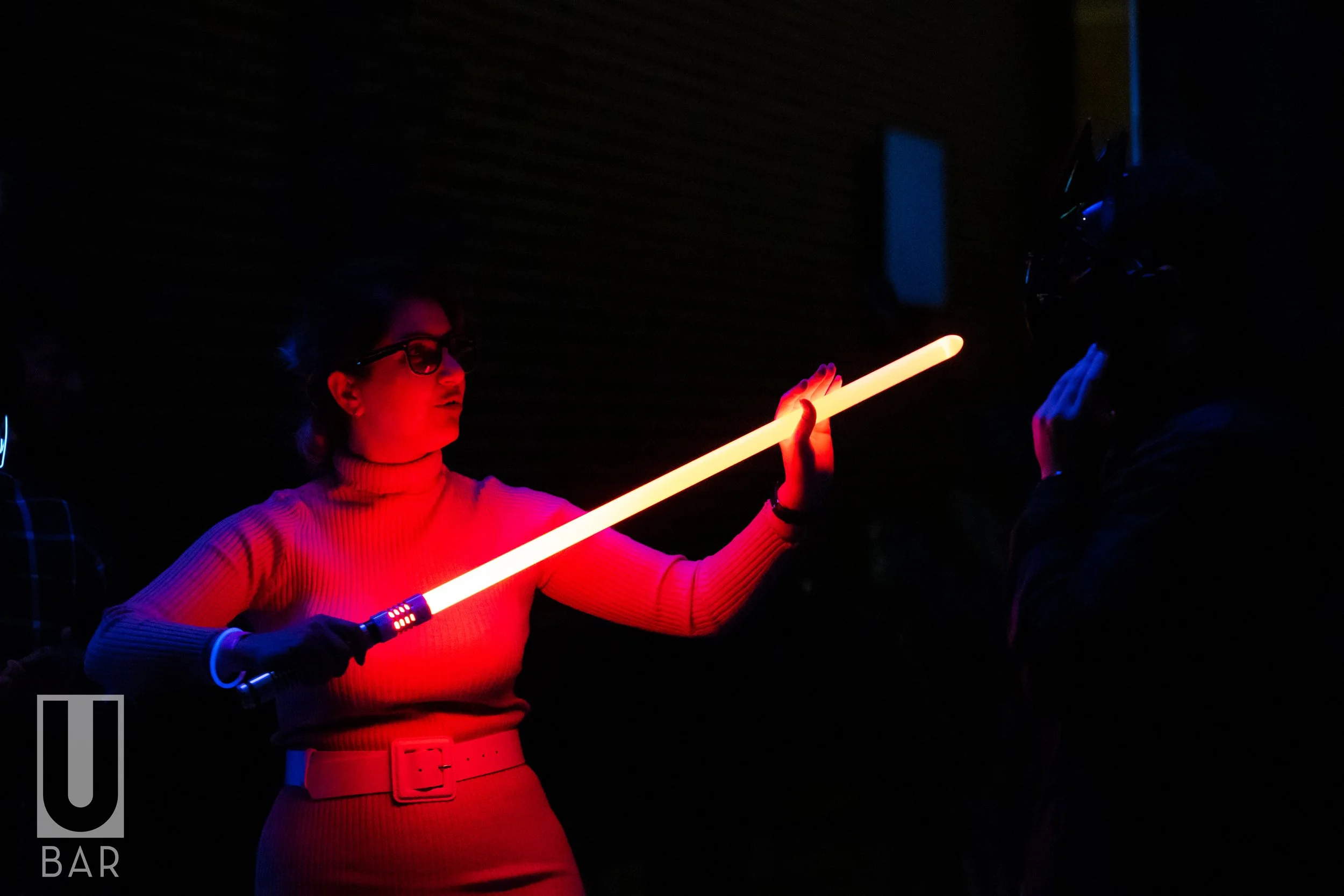 Woman with glasses in a red turtleneck holding a glowing red and yellow light stick, engaging in a light saber duel with a person wearing a dark outfit and helmet inside a dark room.