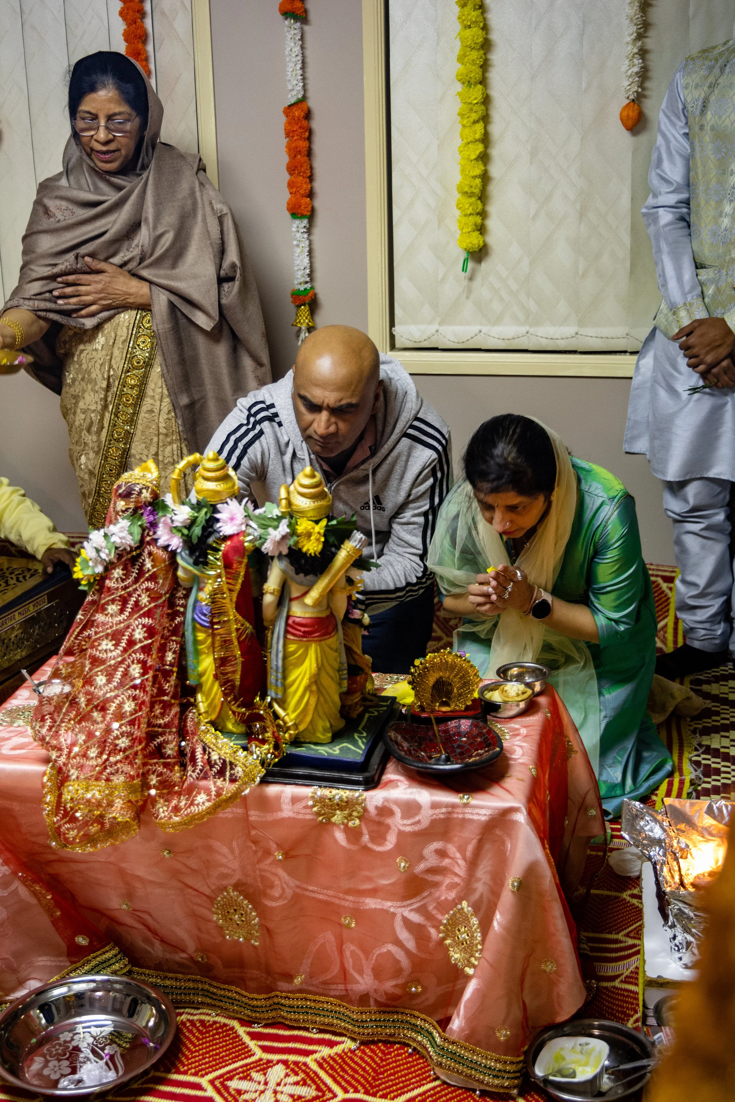 People performing a Hindu prayer or ritual, with a decorated idol of Lord Krishna on a table, surrounded by offerings and traditional items, in a room decorated with marigold flower garlands.