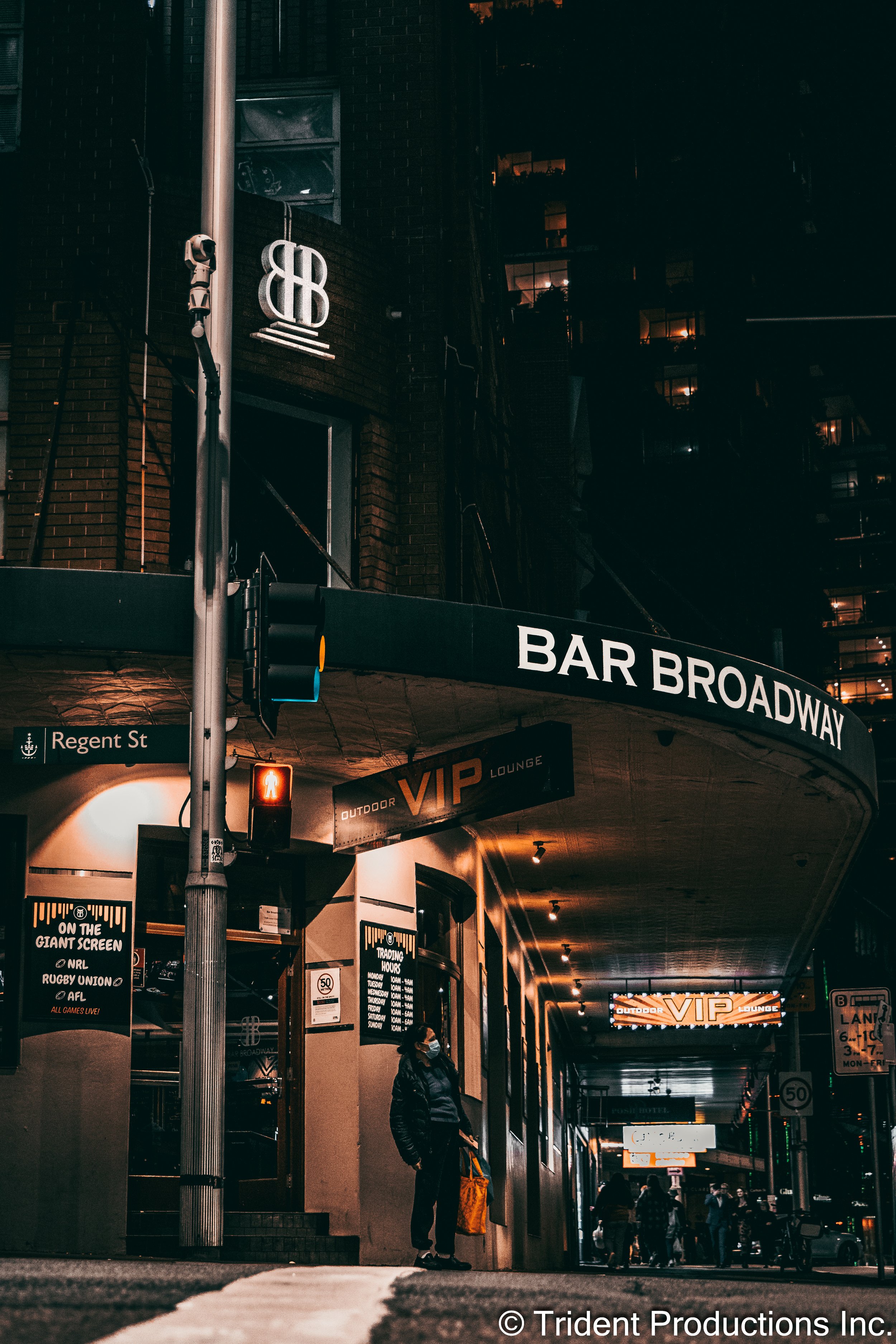 Night view of Bar Broadway with illuminated signs, a woman wearing a face mask standing outside, and people walking in the background on a city street.