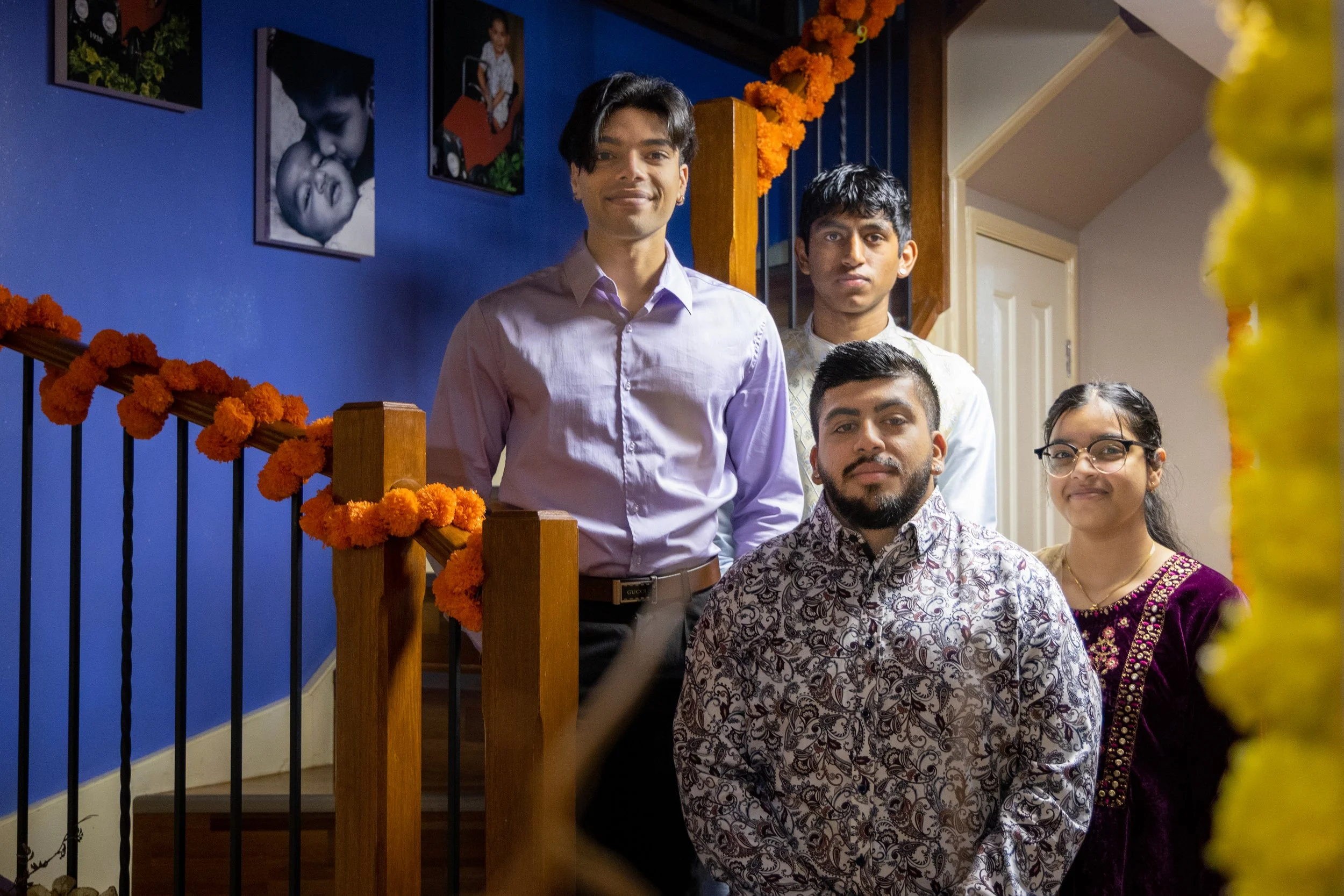 Five people posing indoors during a celebration, with decorations such as marigold flowers and photographs on a blue wall in the background.