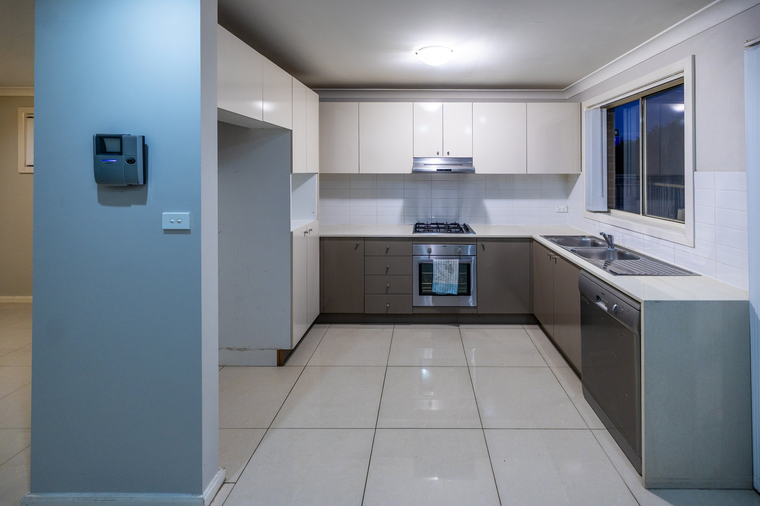 Empty kitchen with beige and white cabinets, a window, a sink, a dishwasher, and a gas stove with an oven. The kitchen has tiled flooring and white tiled walls.