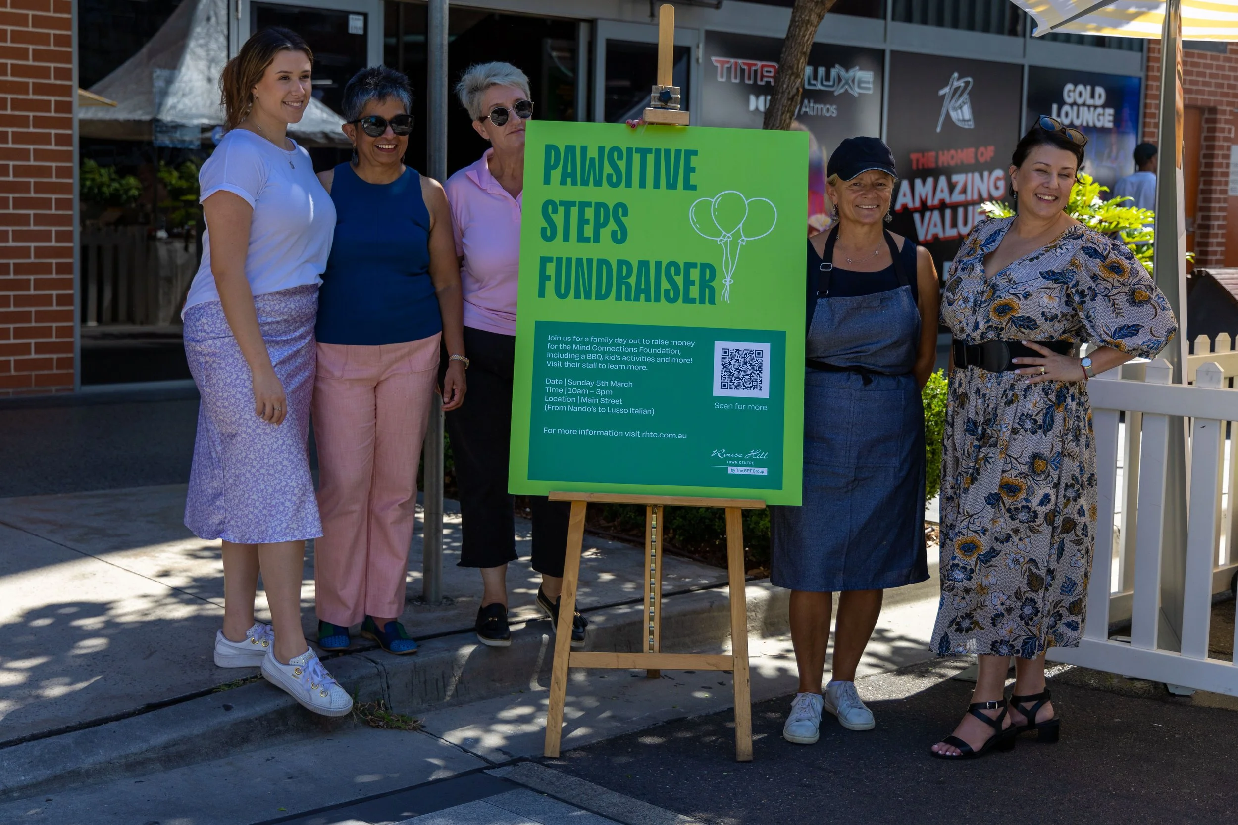 Group of five women standing next to a bright green sign for a fundraiser called 'Pawsitive Steps Fundraiser'. The sign includes event details and a QR code. The women are outdoors, smiling, near a brick building and a white fence.