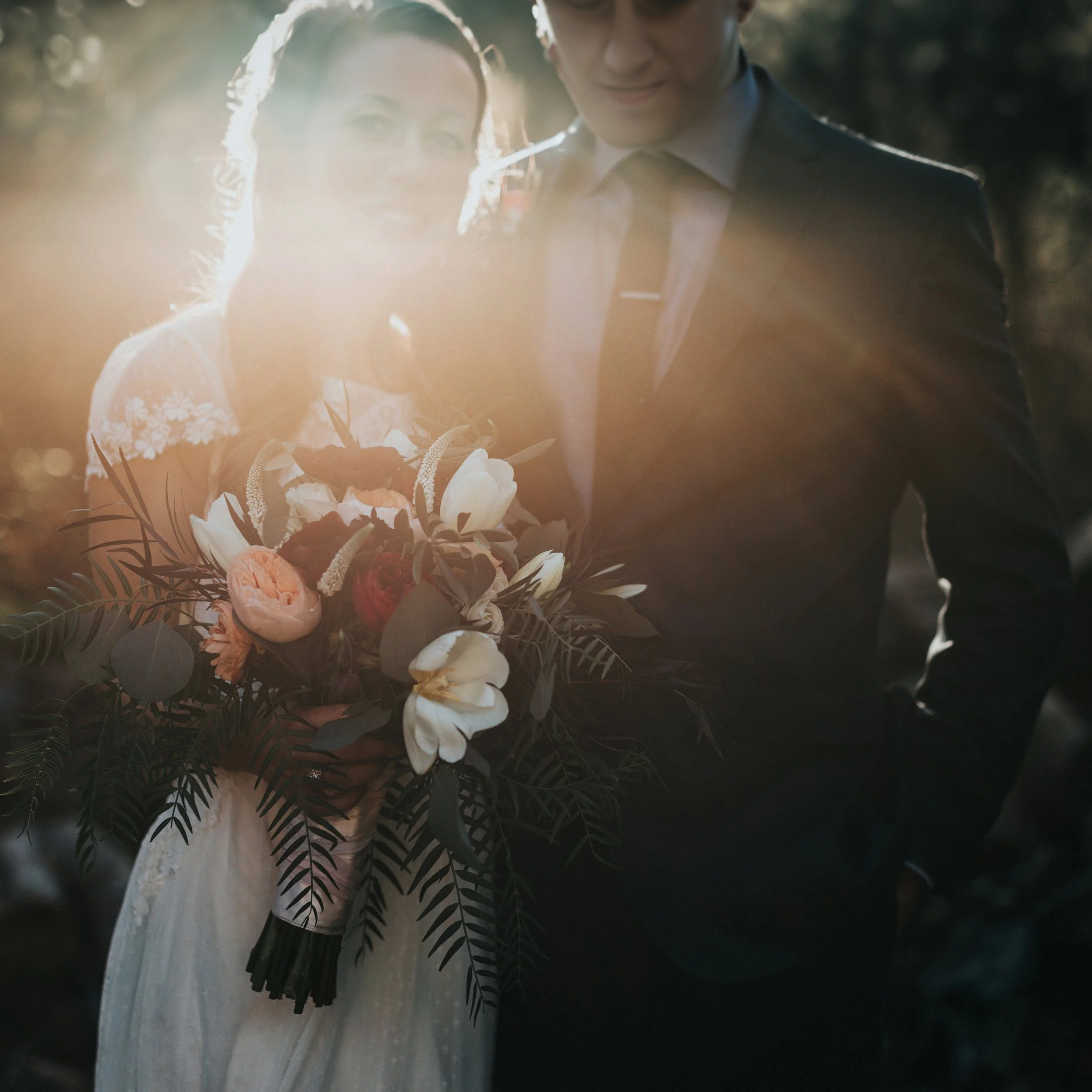 A couple dressed in wedding attire standing close together outdoors during sunset, with the bride holding a bouquet of flowers.