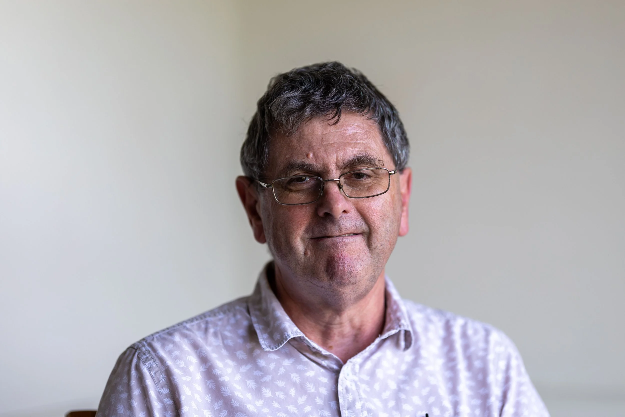 A middle-aged man with glasses, dark wavy hair, and a light-colored patterned shirt, smiling slightly against a plain light background.