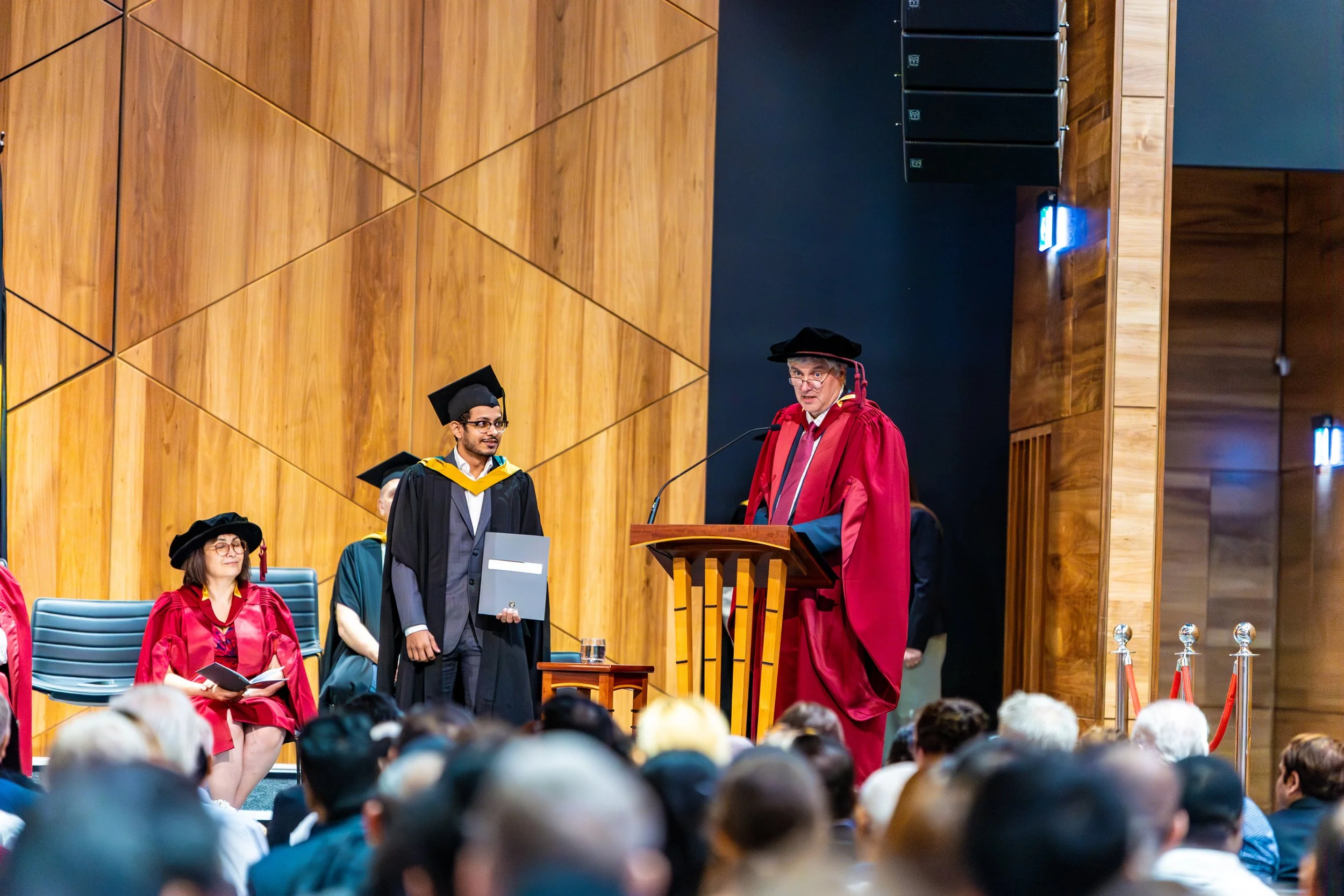 A graduation ceremony with a speaker at a wooden podium and graduates in academic caps and gowns on stage, with an audience in the foreground.