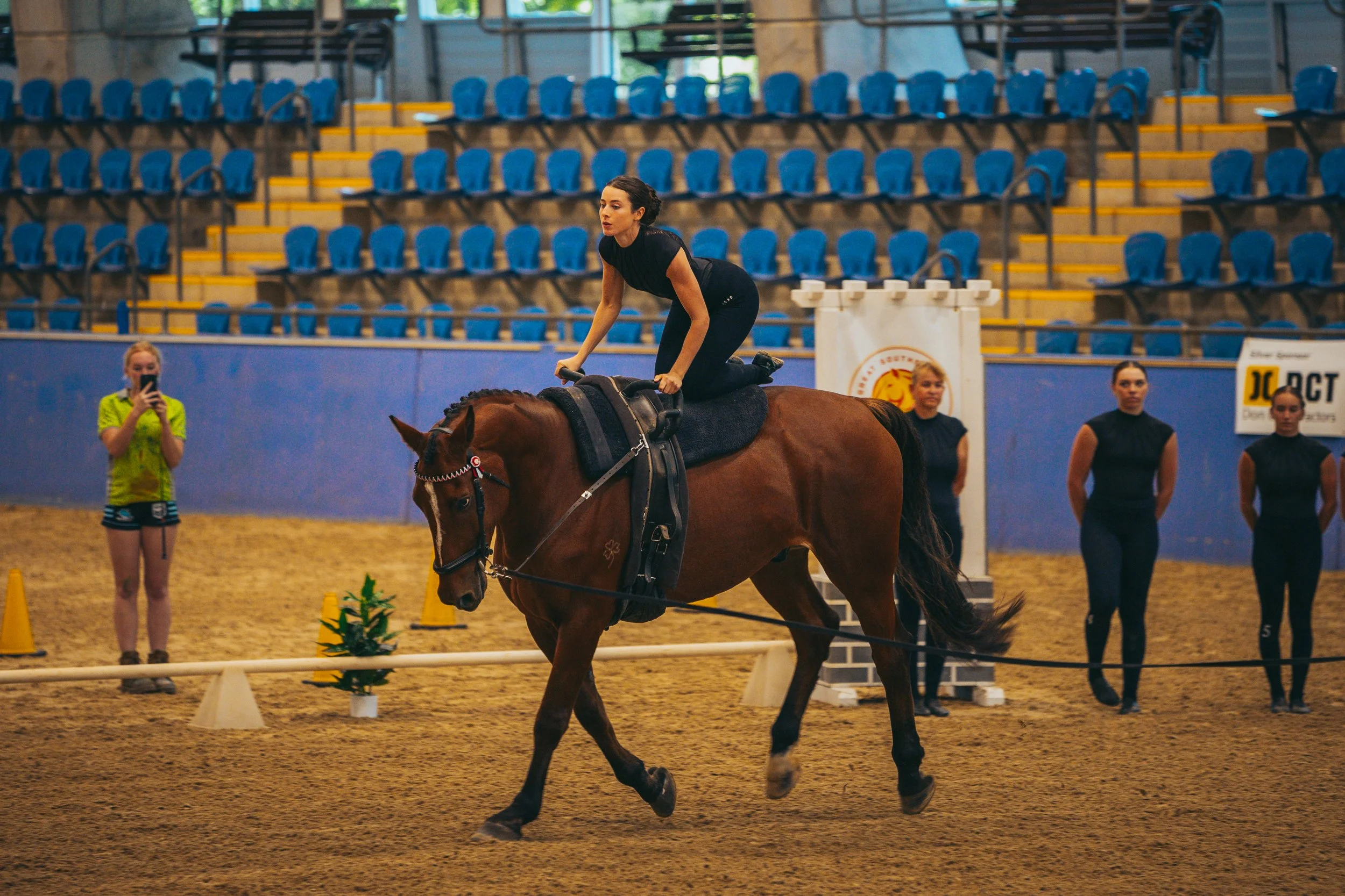A woman riding a horse indoors during an equestrian event. Several spectators and participants are in the background.