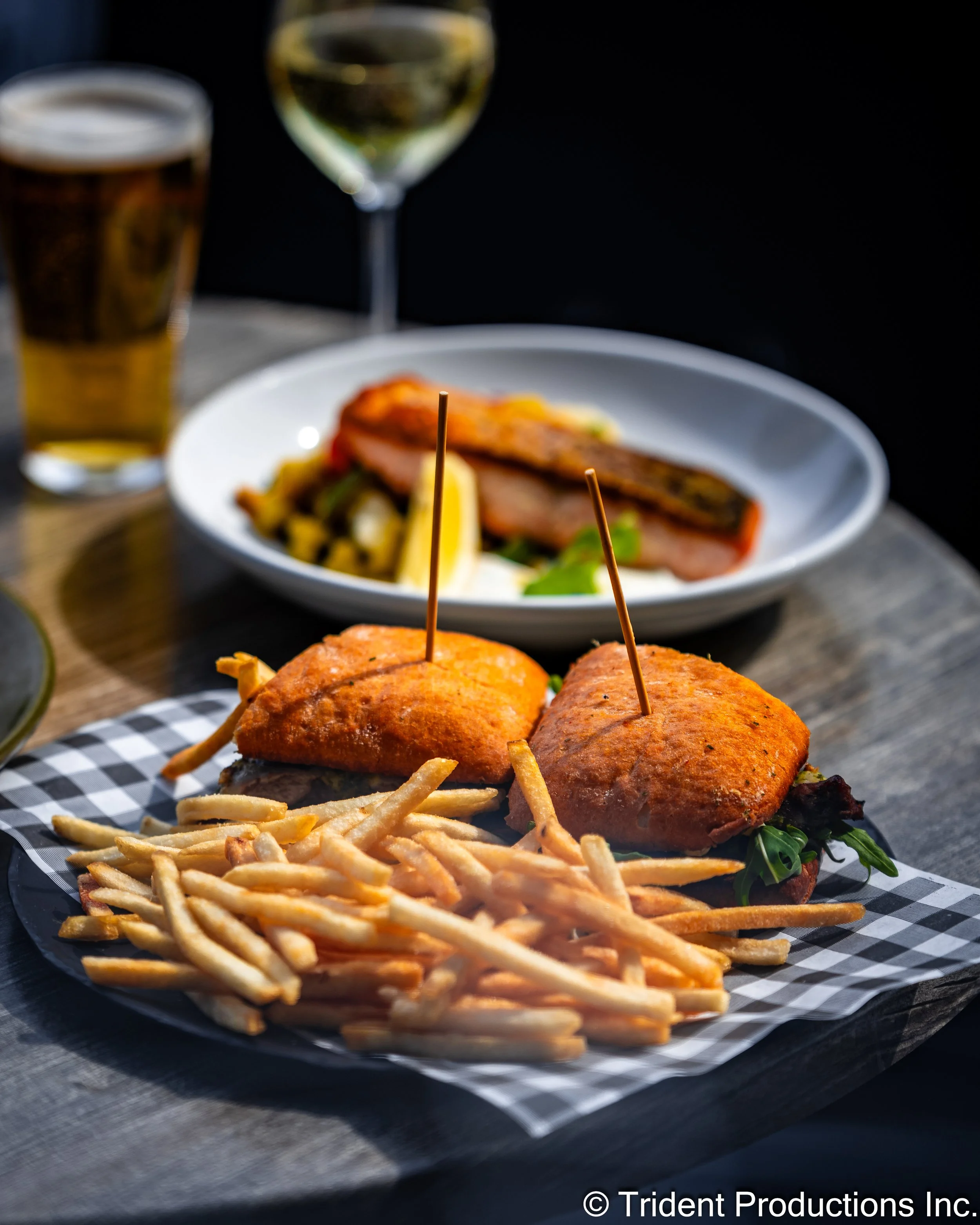 Fried chicken sandwiches with French fries on a checkered paper, served on a black plate in the foreground, with a plate of grilled salmon and salad, and glasses of beer and white wine on a wooden table in the background.
