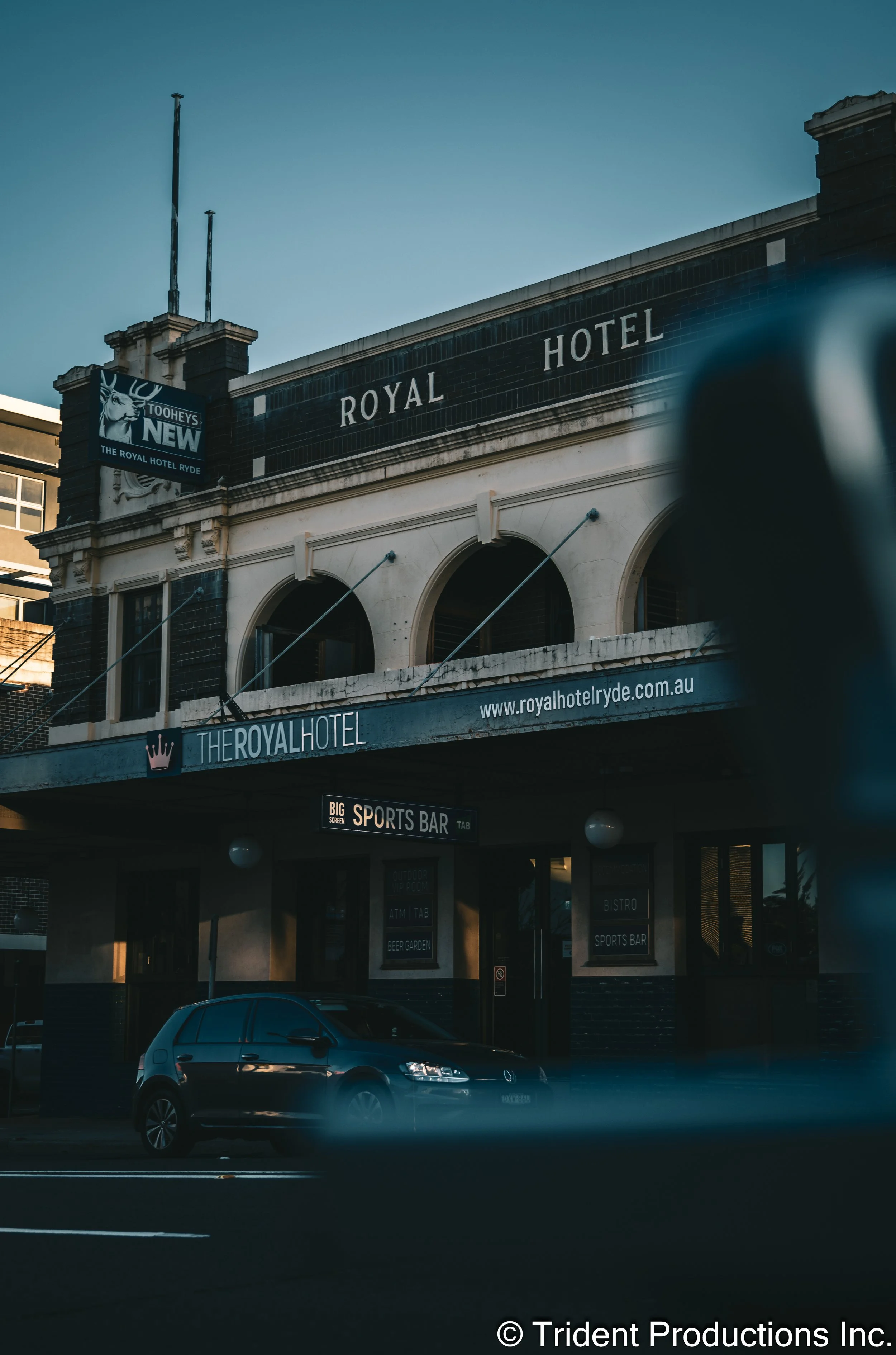The Royal Hotel, a historic building with a sign reading 'Royal Hotel' and a website address, located on a city street with a parked car in front, during late afternoon or early evening.