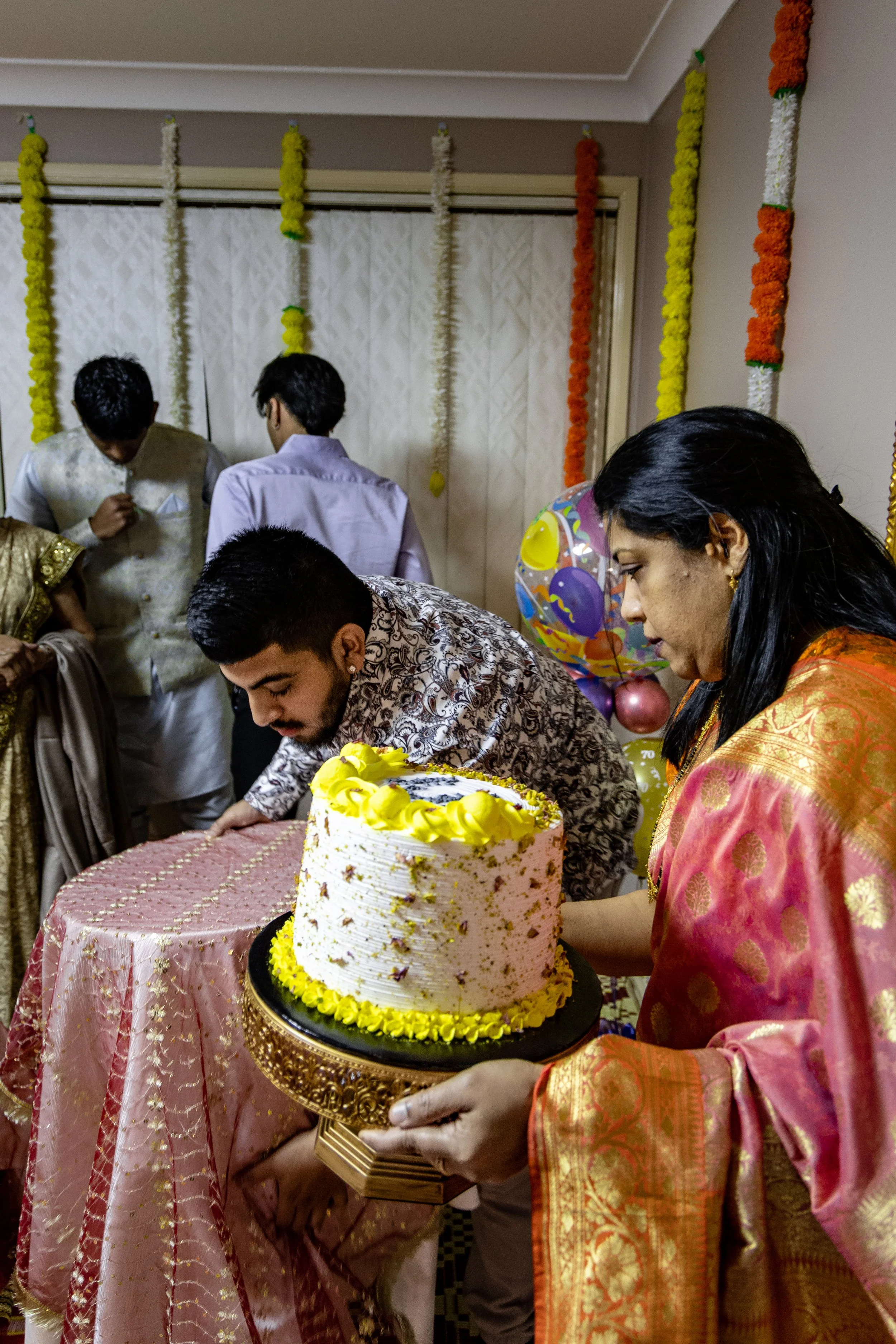 People celebrating a birthday with a decorated cake, balloons, and colorful garlands in the background.