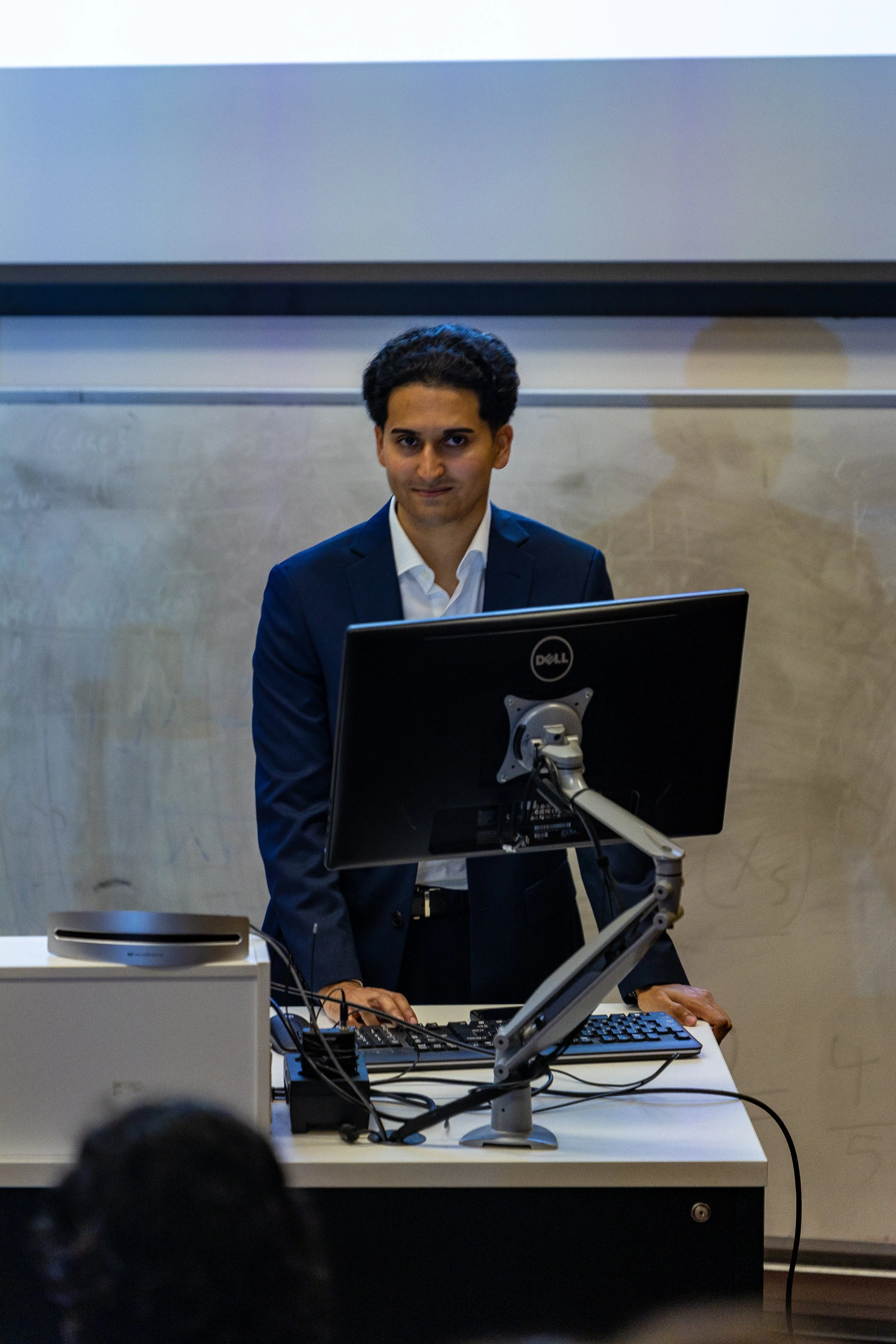 A man in a dark suit standing behind a desk with a computer monitor in a classroom or conference room.
