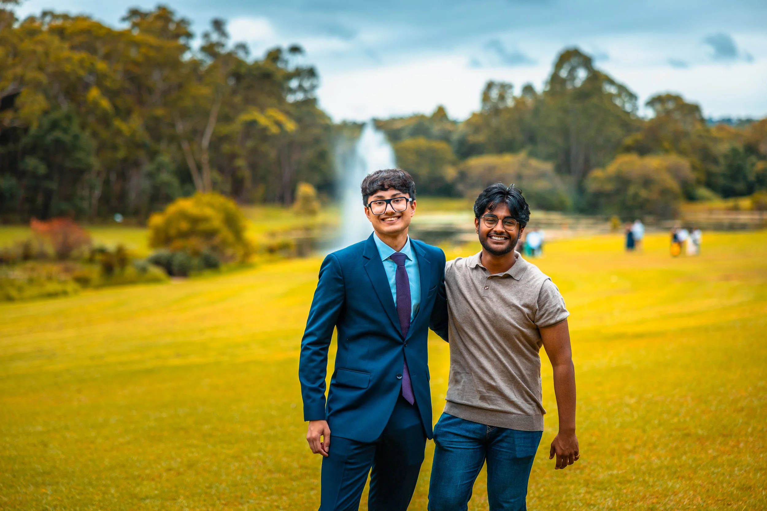 Two young men smiling and posing together outdoors in a park with a fountain and trees in the background.