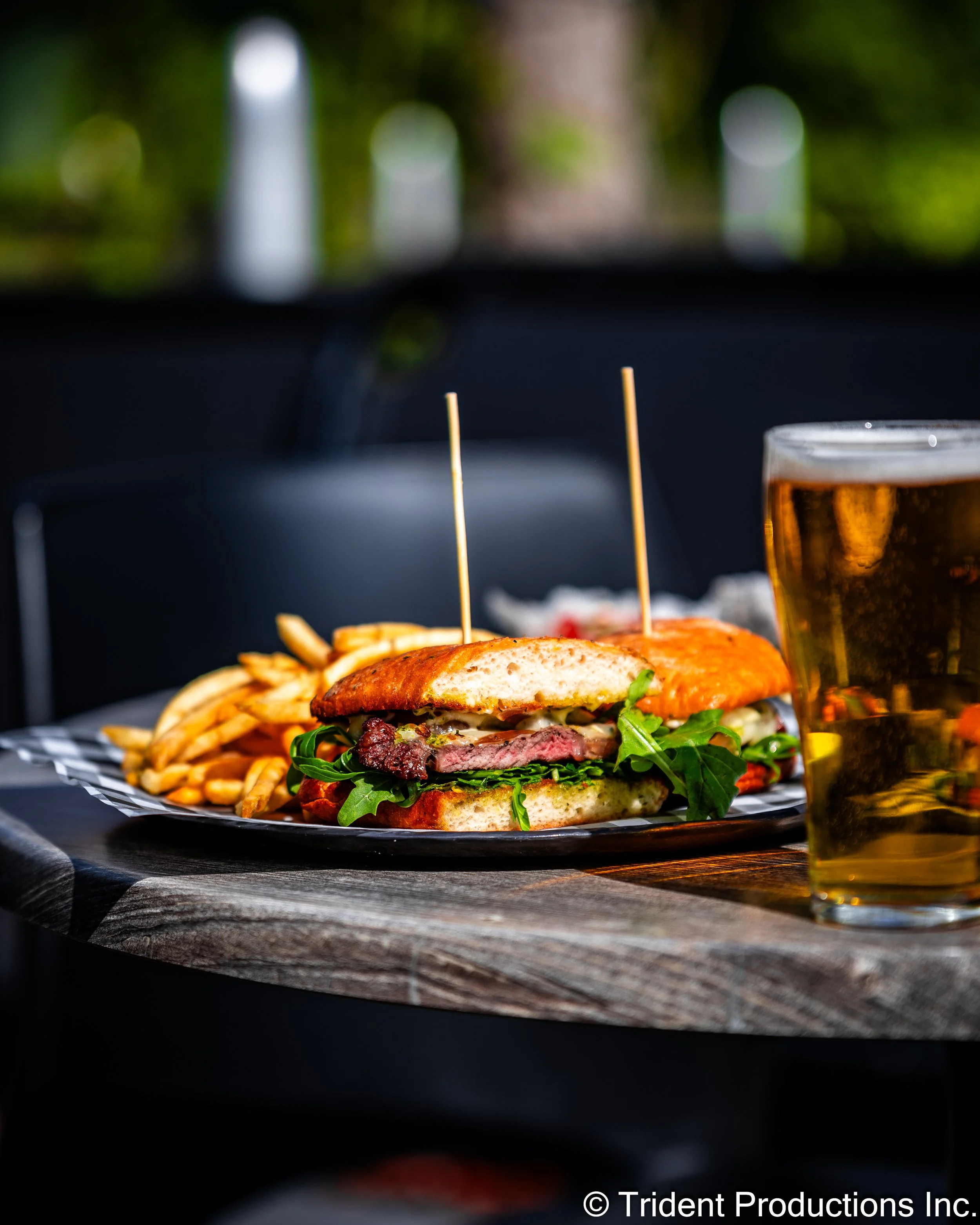 A sandwich with lettuce, tomato, and melted cheese on toasted bread, served with French fries, alongside a glass of beer on a wooden table.