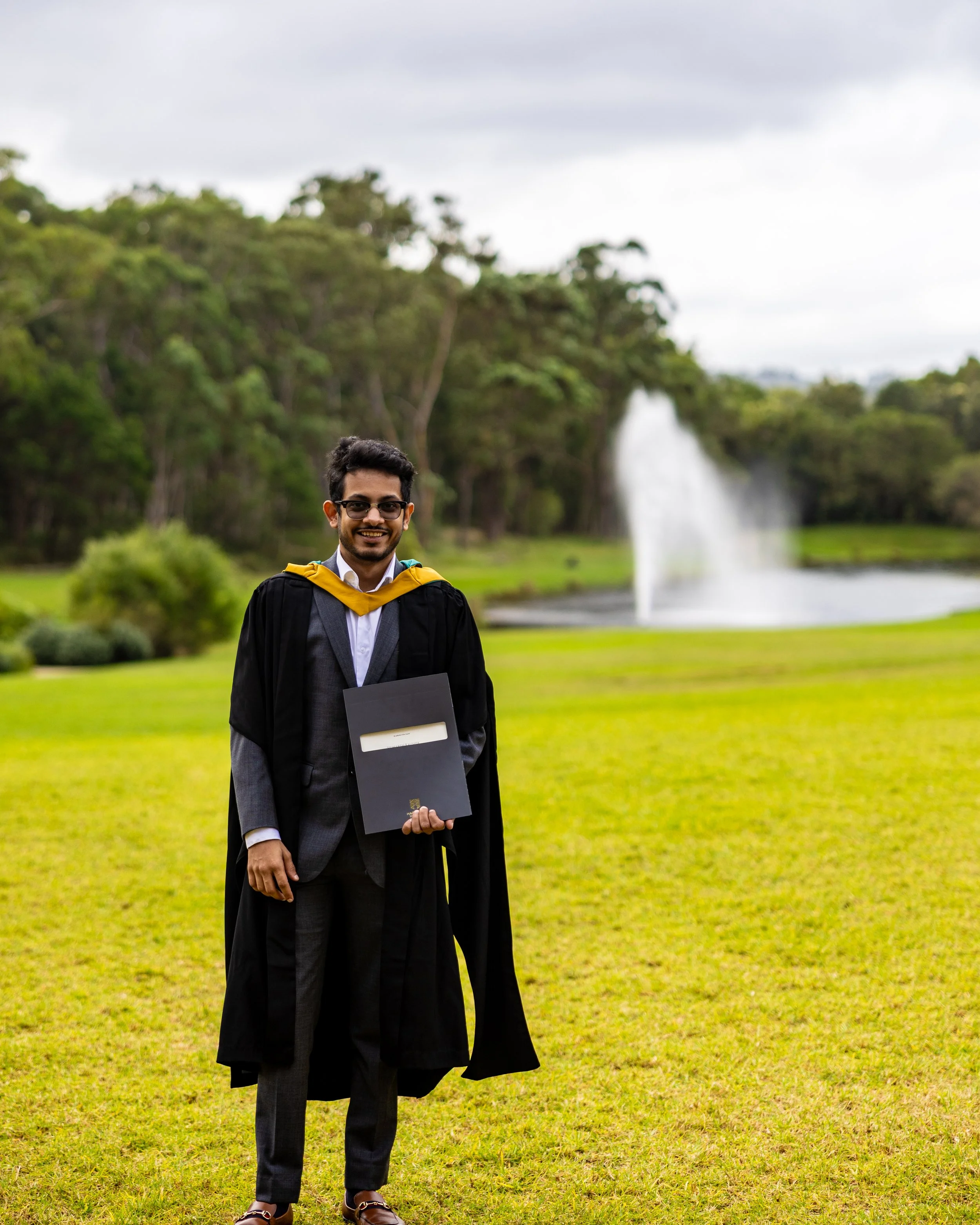A smiling man in a graduation gown and cap holding a diploma, standing on a lush green lawn with trees and a fountain in the background.