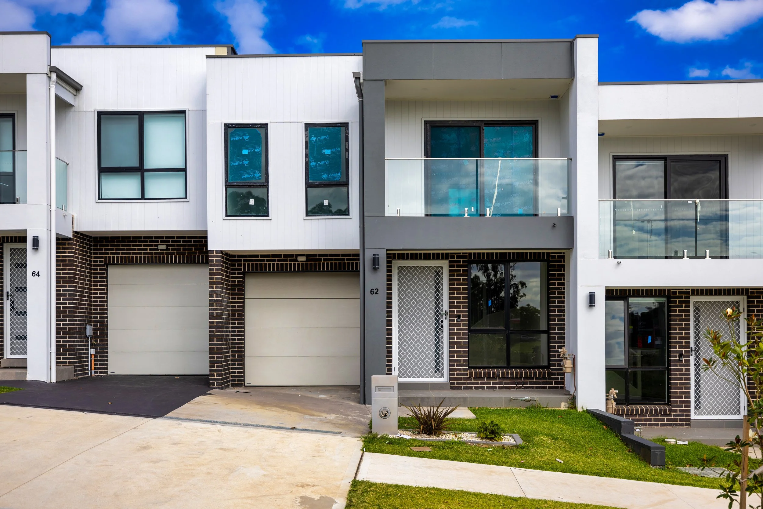 Front view of modern townhouse with brick and white panel facade, two garage doors, and a small garden with grass and plants, under a partly cloudy sky.