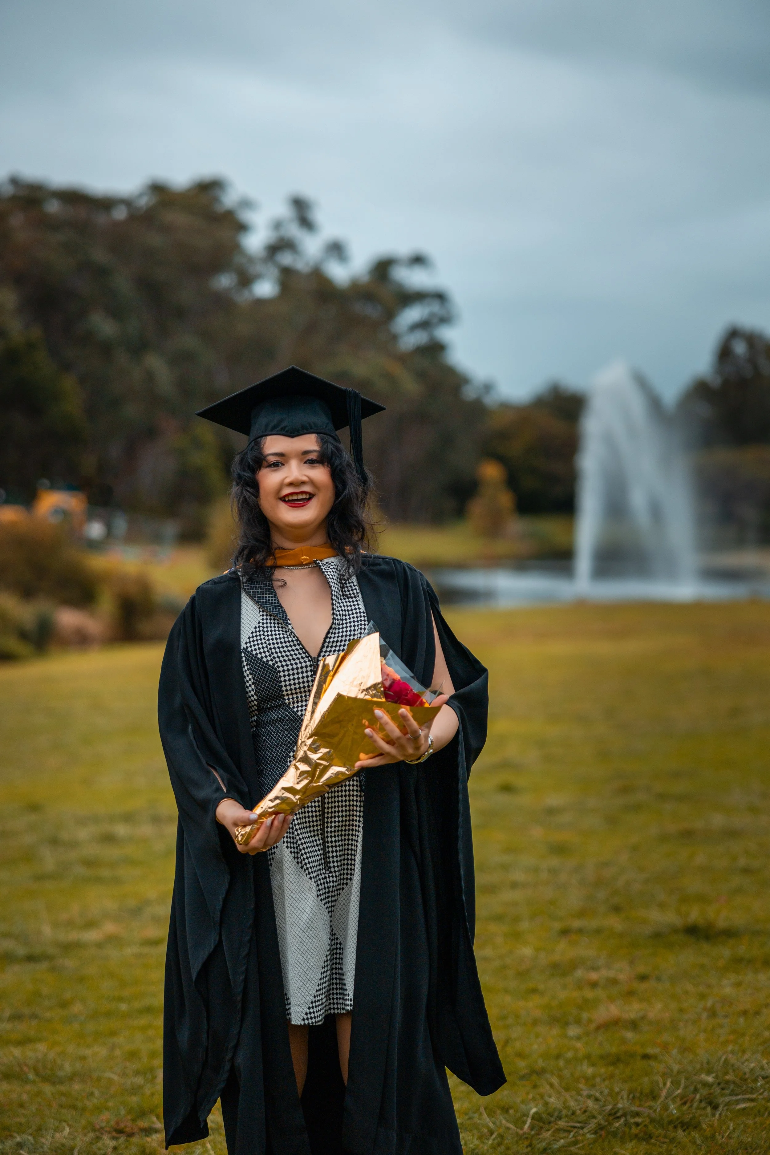A woman in a graduation cap and gown holding a bouquet of flowers standing outdoors near a fountain and trees.
