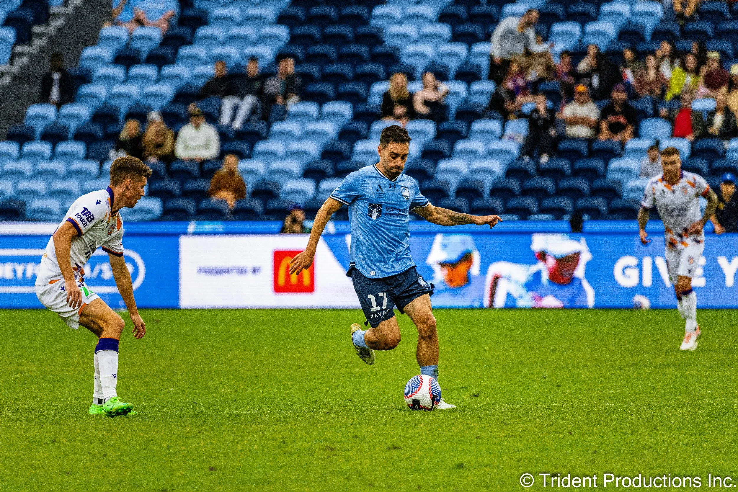 A soccer player in a blue uniform preparing to kick a ball during a game on a grassy field. Two other players in white uniforms are nearby, and spectators are seated in a mostly empty stadium in the background.