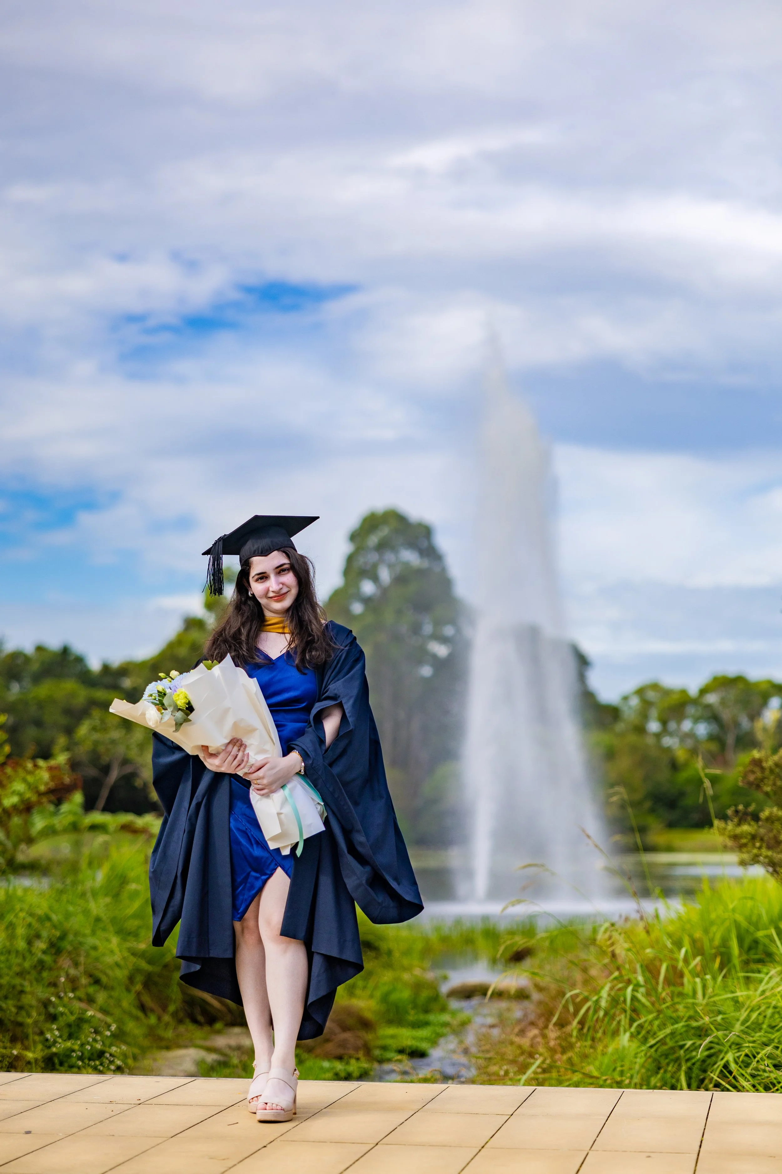 A young woman in graduation cap and gown holding a bouquet of flowers, standing on a wooden pathway outdoors with a fountain and trees in the background.
