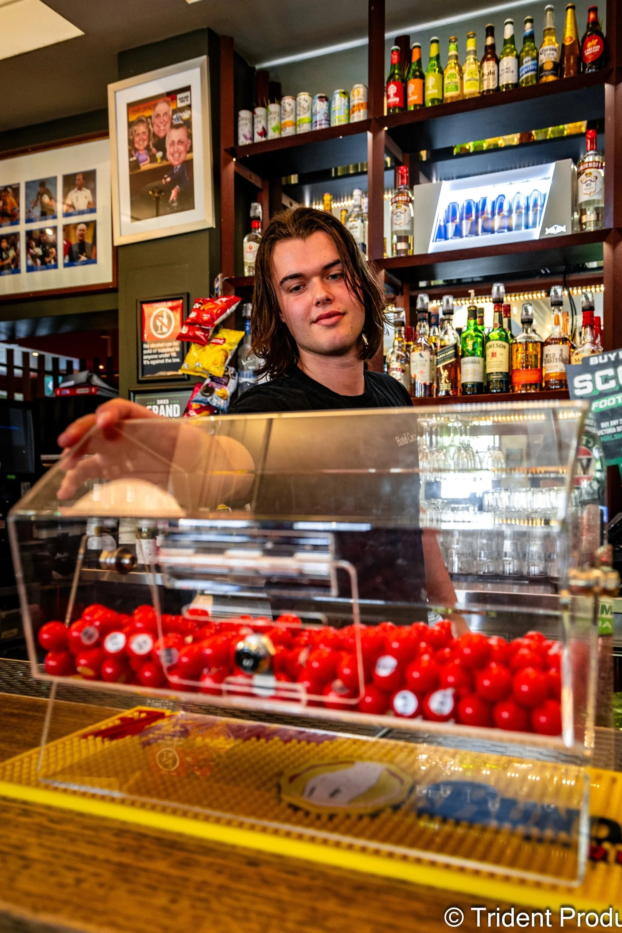 Young man standing behind a counter at a bar, with rows of liquor bottles behind him, and a display case of red cherries in the foreground.