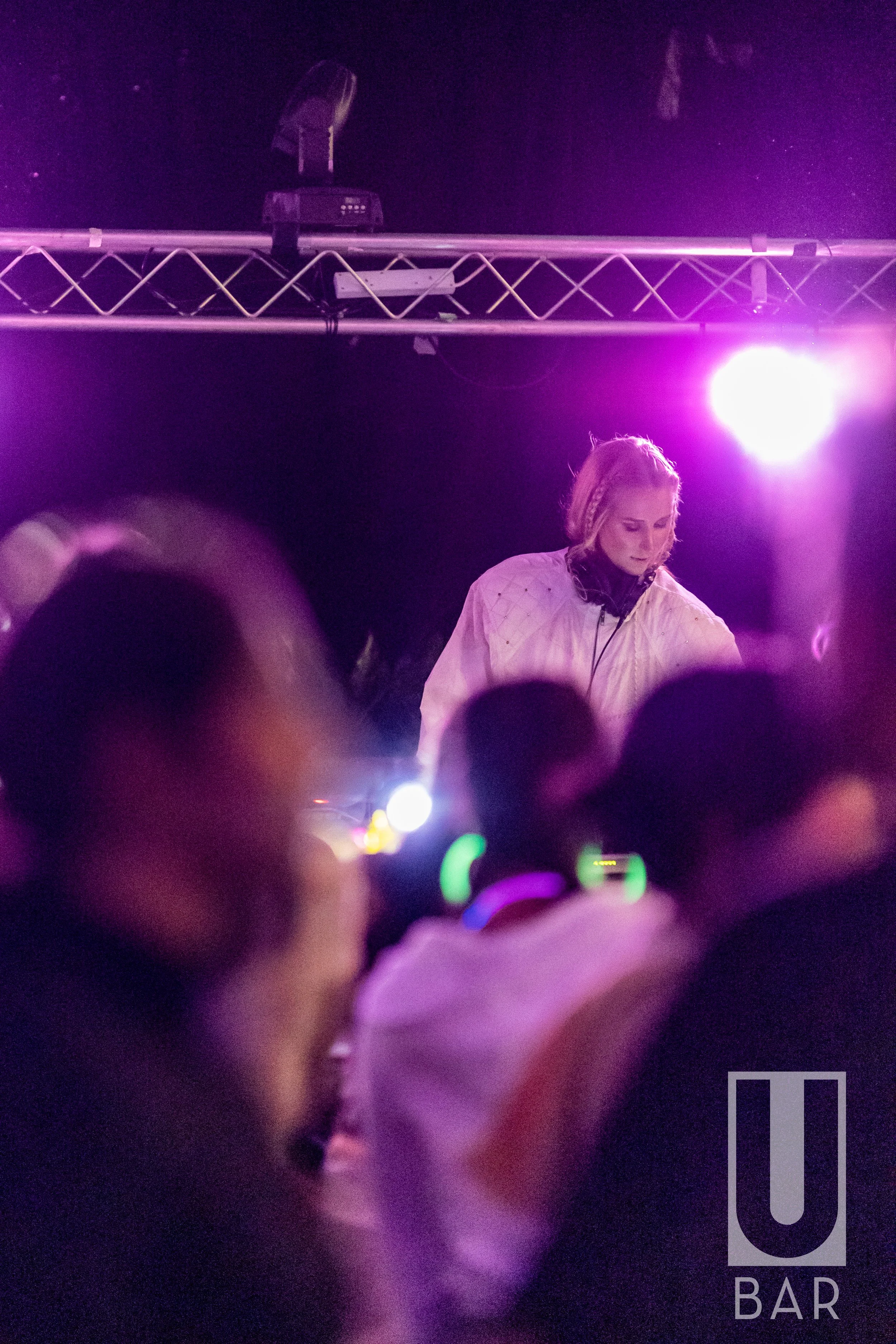 A female DJ wearing a white jacket and headphones performing at a nightclub with purple lighting, with blurred crowd members in the foreground and a DJ booth visible.