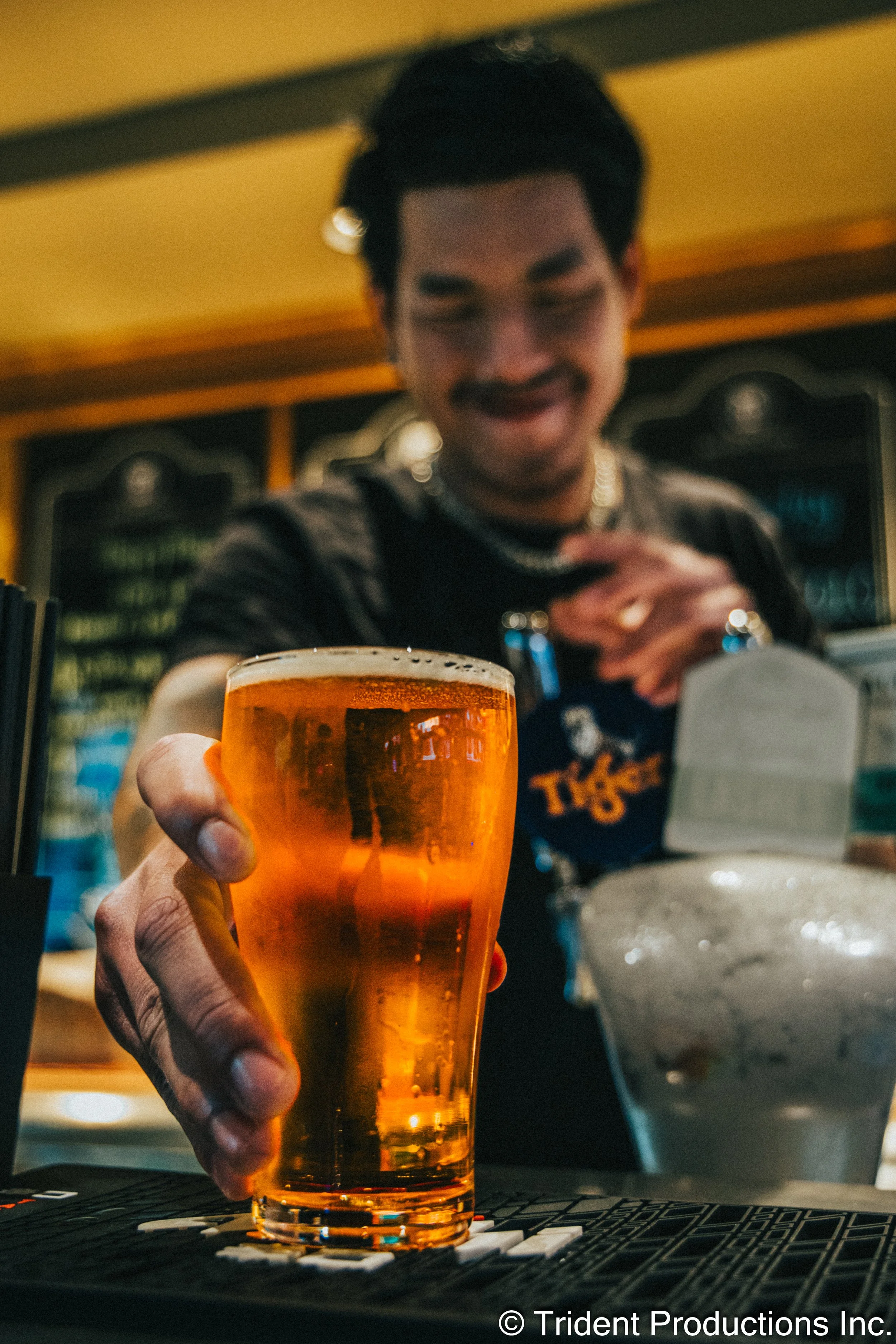A bartender presenting a pint of beer with a smile, with a chalkboard menu in the background.
