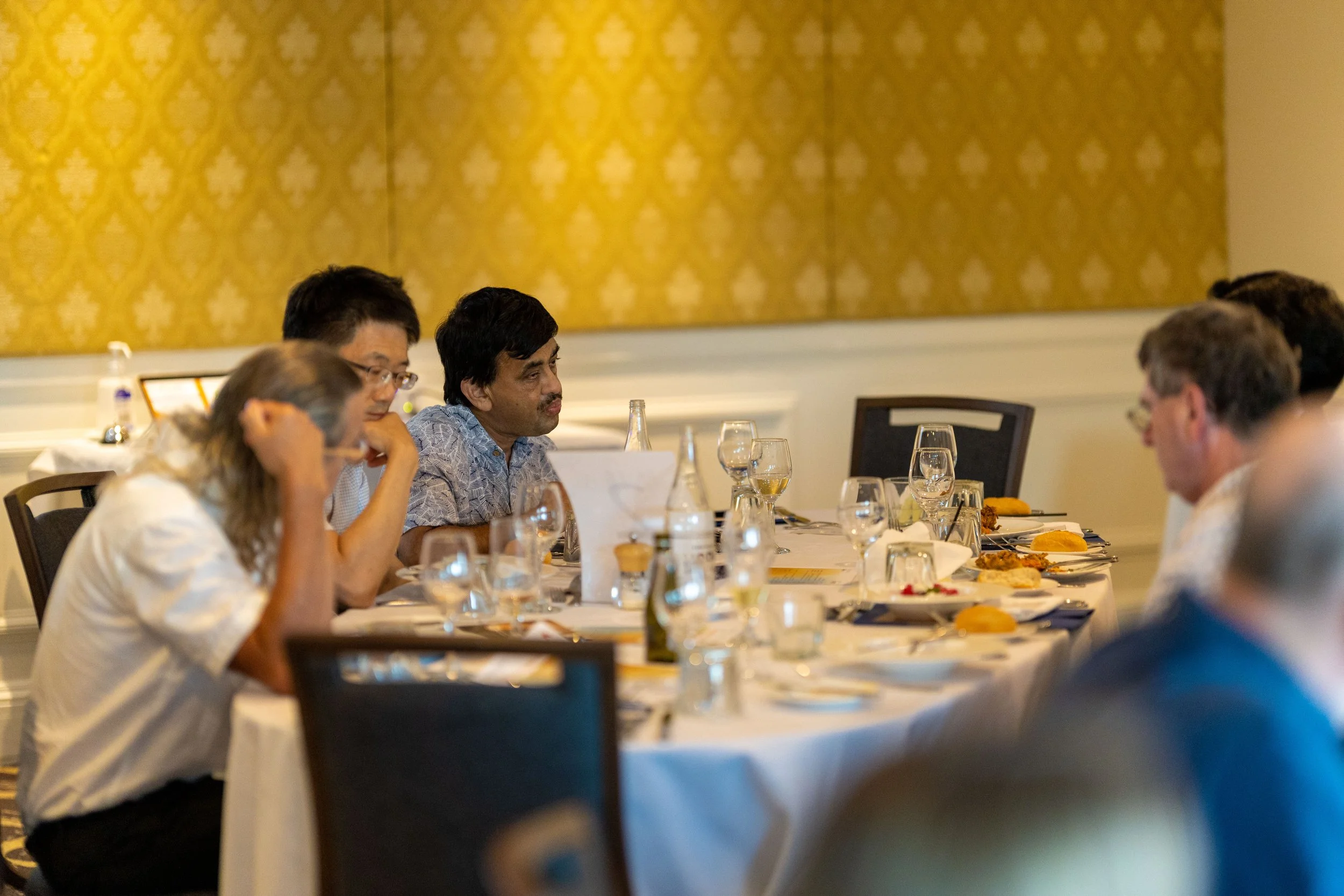 People sitting at a round dining table in a formal setting, engaged in conversation, with plates of food, glasses, and bottles on the table.