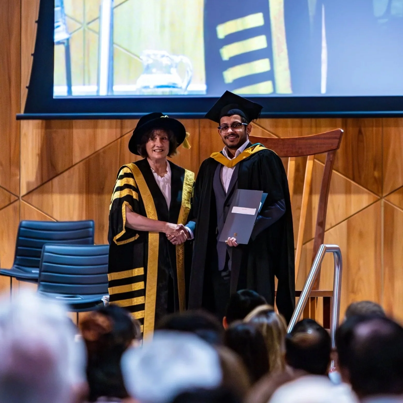 A young man in graduation cap and gown receiving a diploma from a woman in academic regalia during a graduation ceremony.