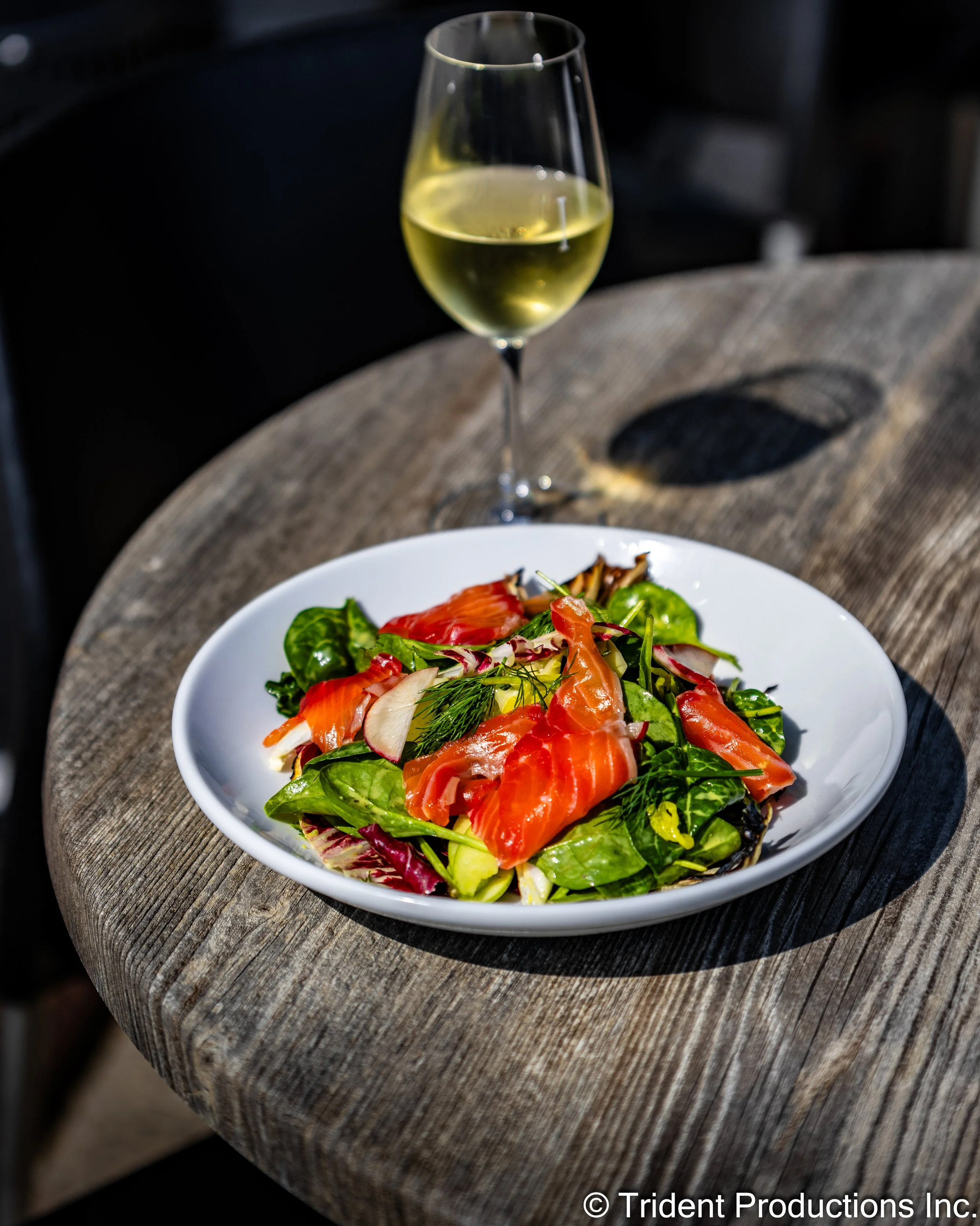 A plate of mixed salad with smoked salmon, leafy greens, radish slices, and herbs, with a glass of white wine on a rustic wooden table.