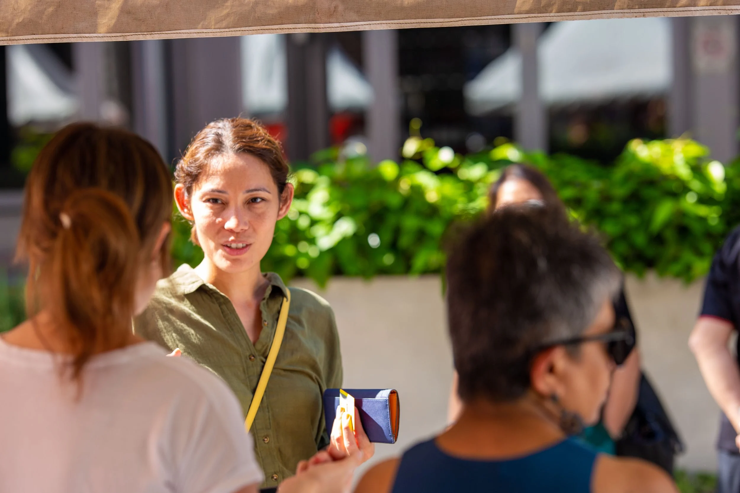Group of people outdoors, woman in green shirt talking to others, one person holding a card, greenery in background.