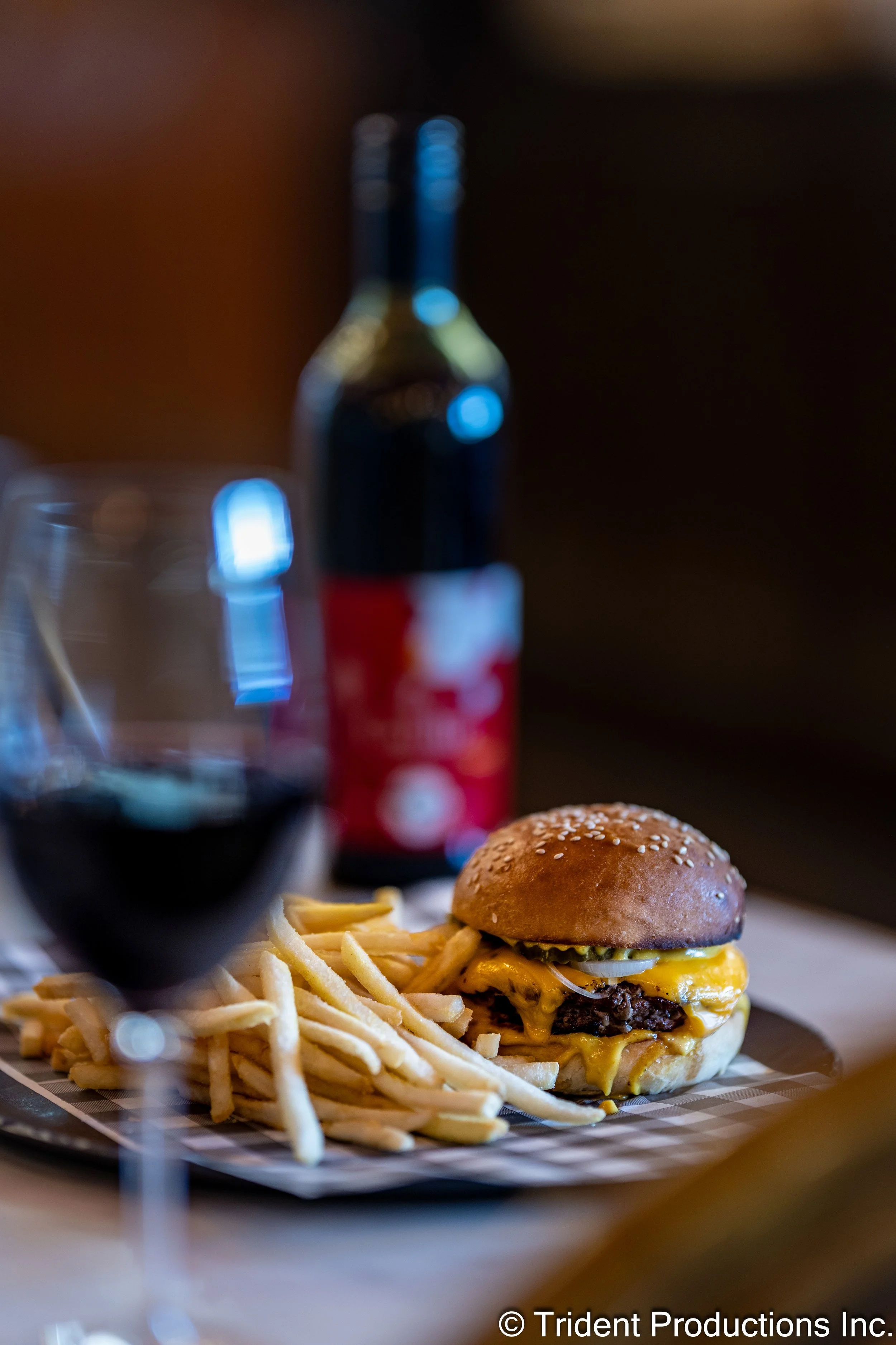 Close-up of a cheeseburger with pickles, onions, and melted cheese, served with French fries. A glass of red wine and a bottle of soda are in the background, on a plate with checkered paper.