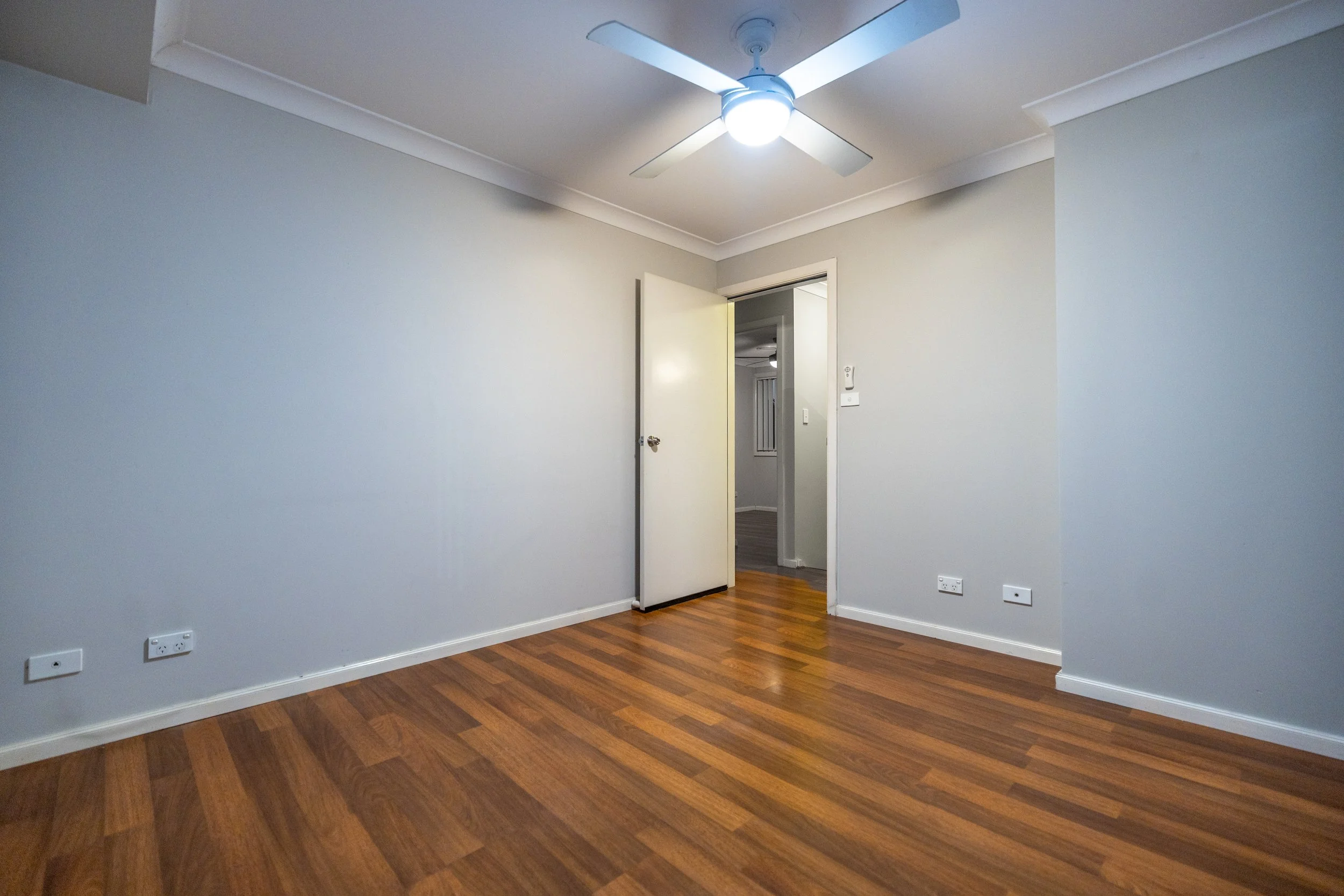 Empty living room with light-colored walls, hardwood floors, a ceiling fan, and an open door leading to another room.