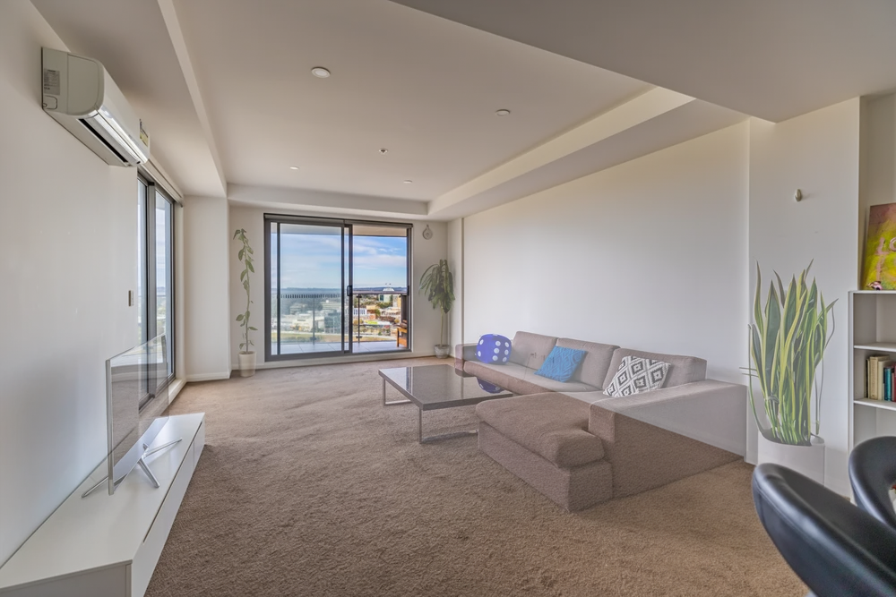Living room with large windows, beige carpet, gray sofa with blue and patterned pillows, glass coffee table, potted plants, air conditioner, and bookshelf.