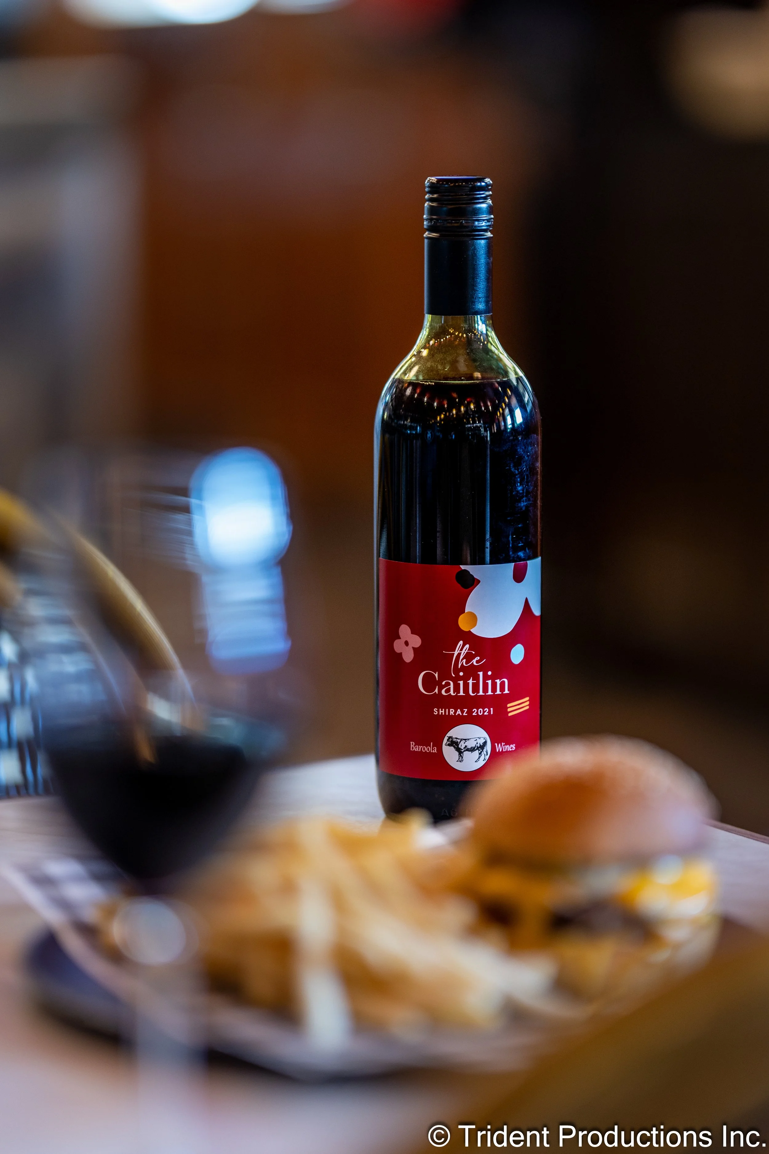 A bottle of red wine labeled 'The Caitlin Shiraz 2021' with a red, white, and black abstract label, placed on a table next to a glass of red wine, some potato chips, and a burger in a blurred background.