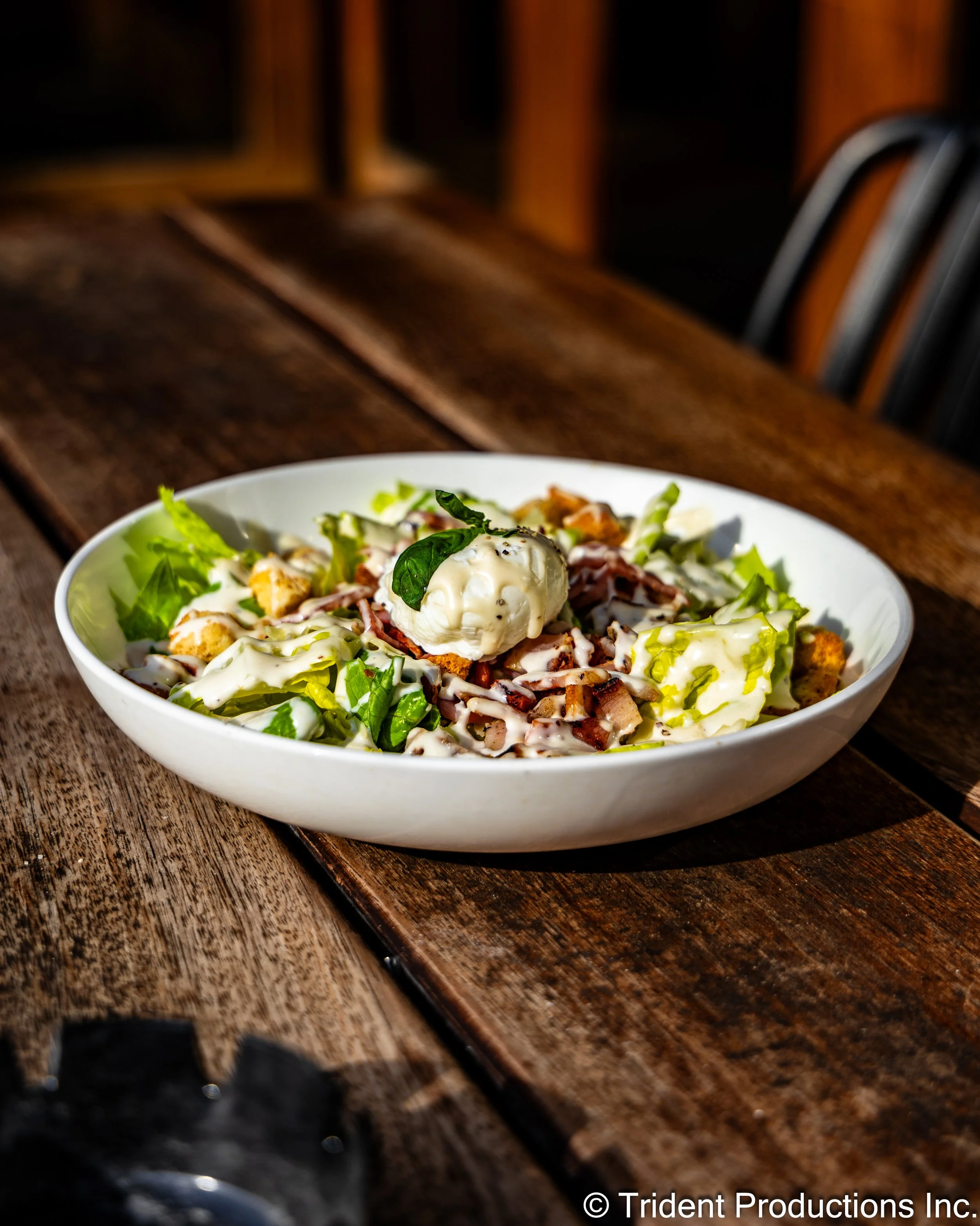 A white bowl filled with a salad containing lettuce, bacon, croutons, a dollop of creamy dressing, and a basil leaf garnish, placed on a wooden table.