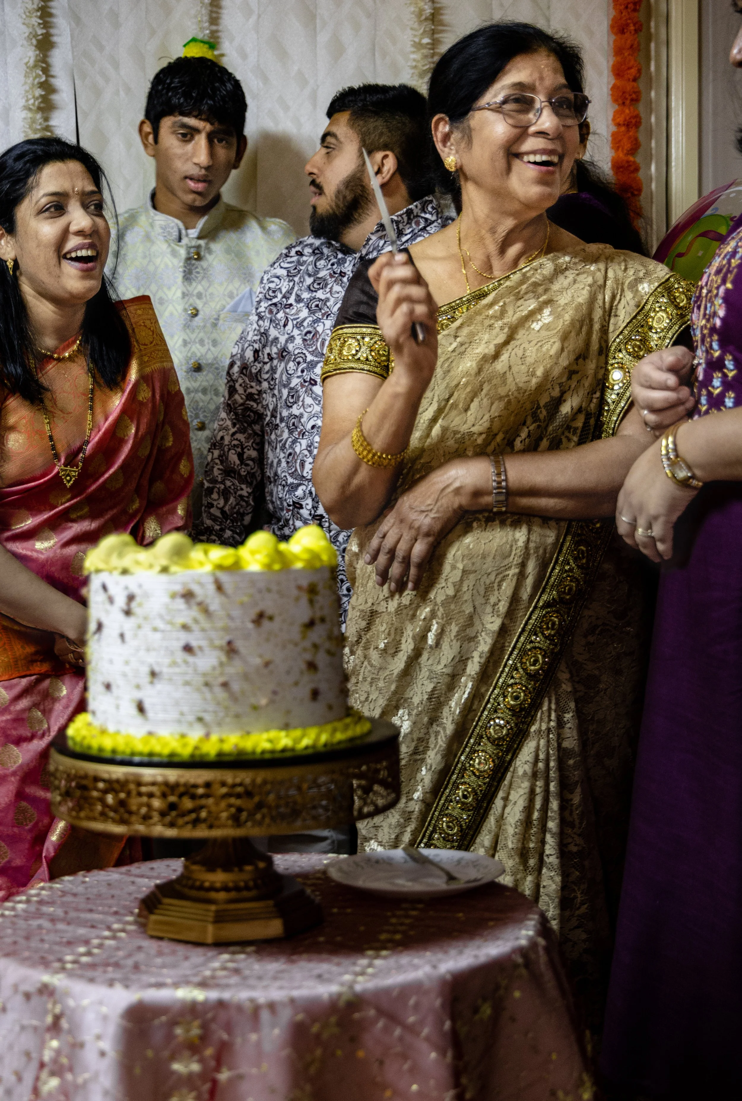 Group of people celebrating around a decorated cake during a festive event, with women wearing traditional Indian sarees and men in ethnic attire.