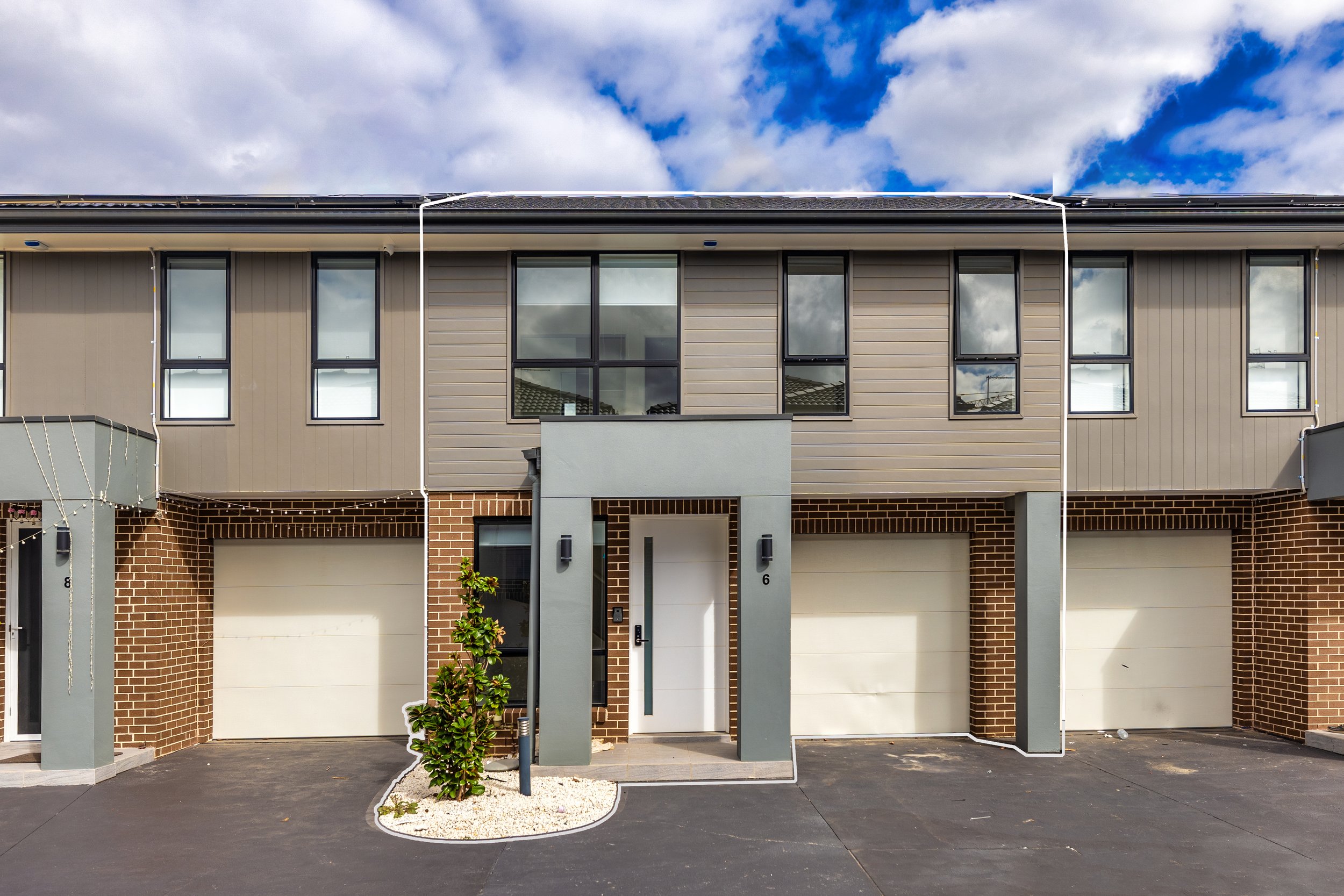 Modern three-story townhouses with brick and siding exteriors, garage doors in the front, and a small landscaped area with a bush and white stones, under a partly cloudy sky.