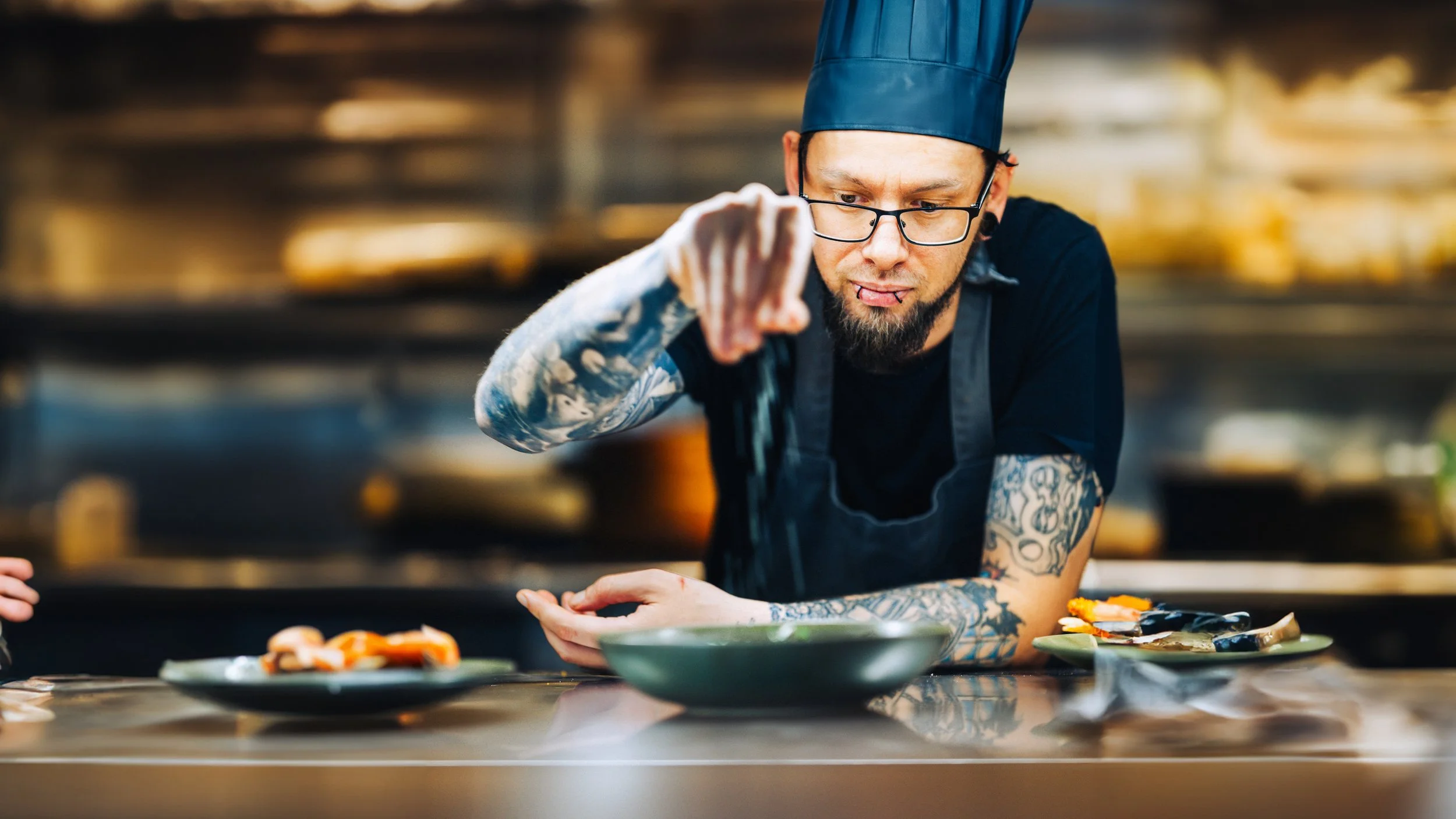A tattooed chef wearing a black apron and a blue chef's hat looks intently at a dish he is preparing in a professional kitchen.