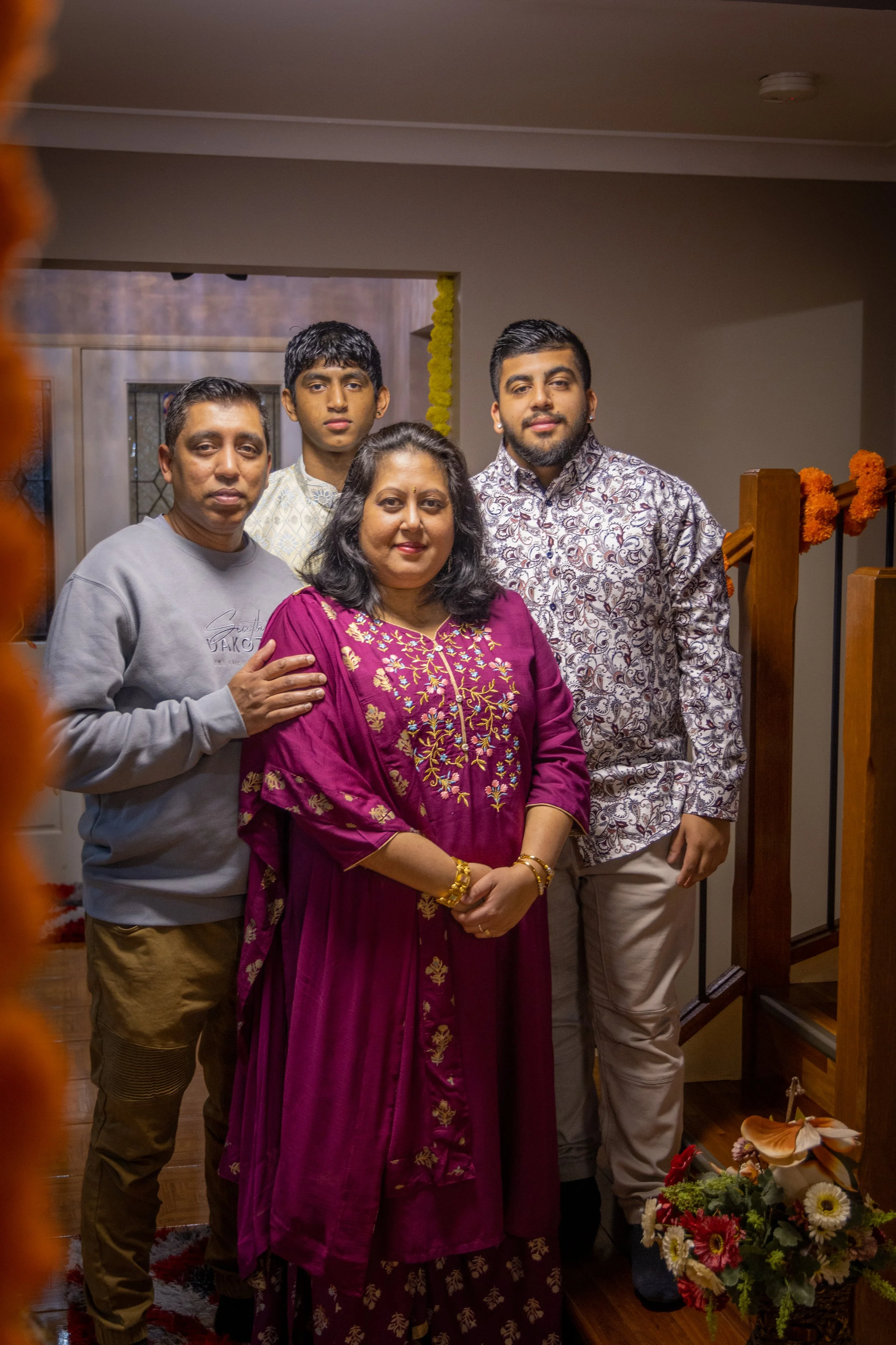 A family of five posing together indoors during a celebration, with festive decorations including orange marigold garlands and a floral arrangement.