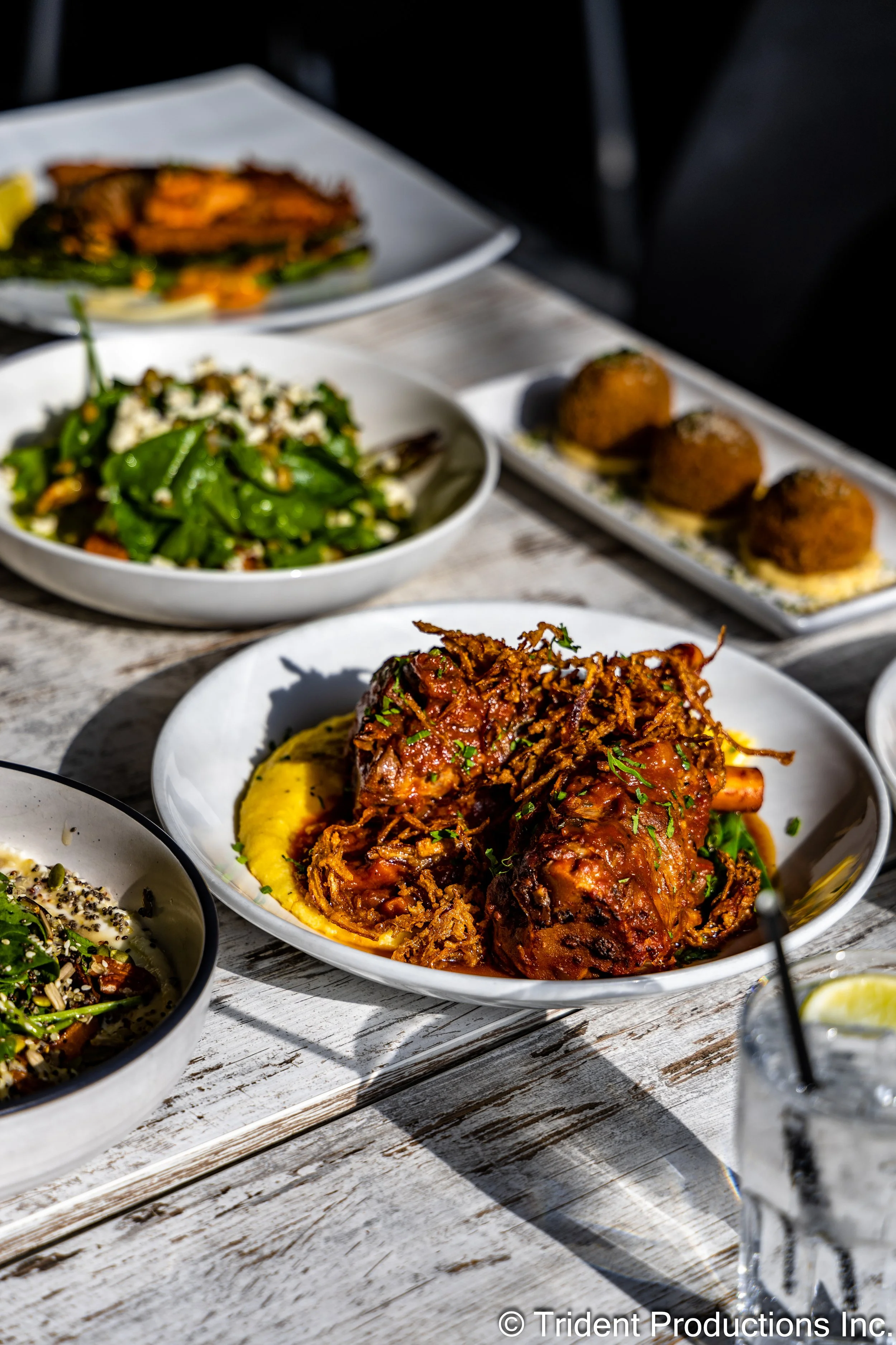 A variety of plated dishes on a rustic wooden table, including braised beef shanks with mashed potatoes, a green salad with vegetables and seeds, croquettes, and a spinach salad with nuts and cheese, with sunlight casting shadows.