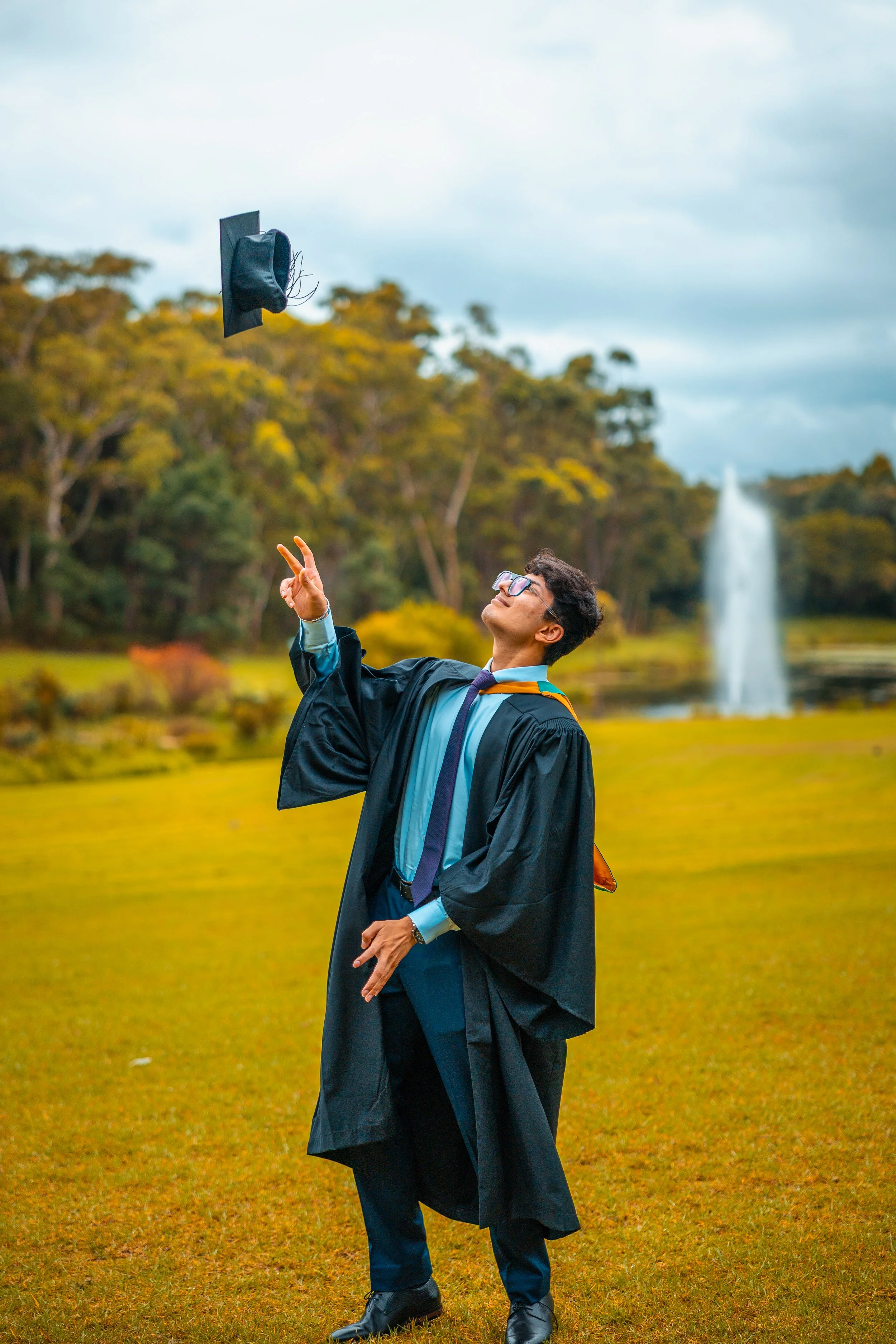 A university graduate in cap and gown tossing his mortarboard in the air outdoors, with a lake, fountain, and trees in the background.